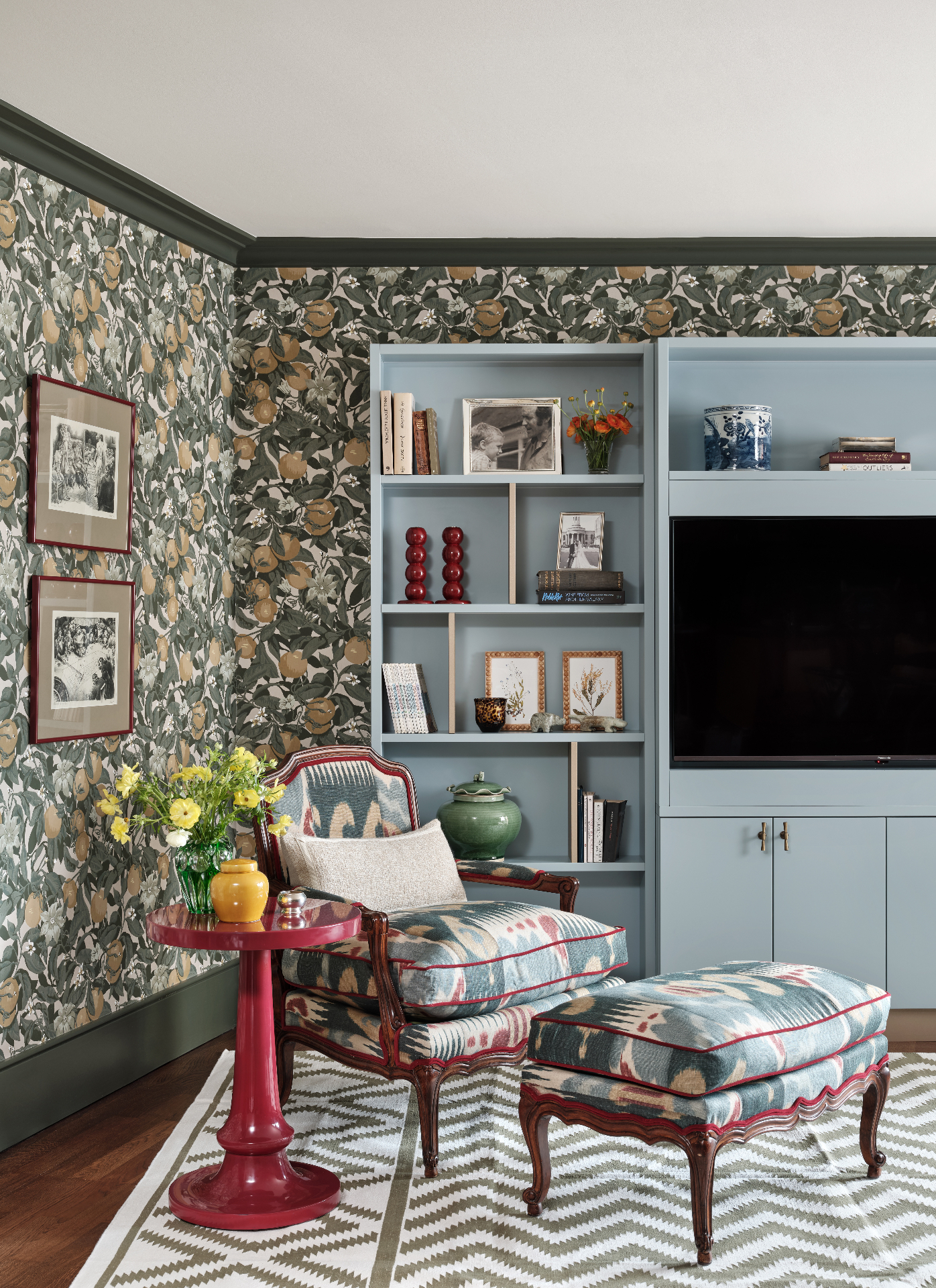 a living room with white green and yellow botanical wallpaper, a red shiny side table and a blue and red patterned armchair with footstool