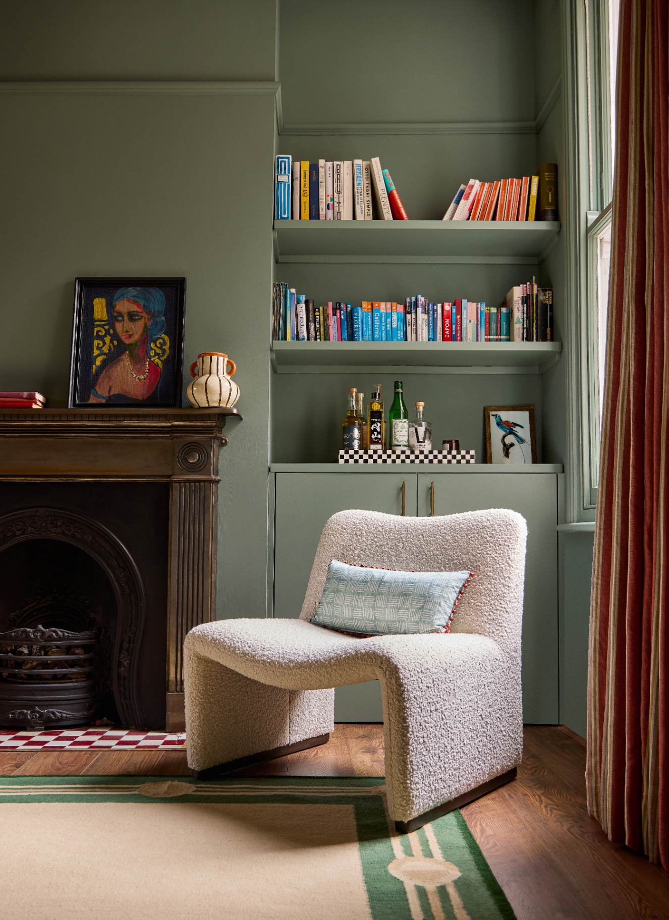 A serene, sage-green living corner featuring a sculptural white bouclé lounge chair, an alcove with built-in bookshelves over a drinks cabinet, a dark wood mantelpiece with framed art, and terracotta striped drapes.