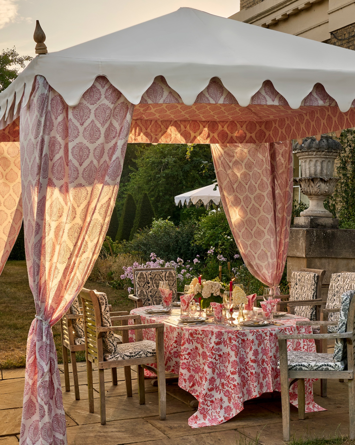 a large cream gazebo wit ha white and pink block printed interior canopy and a scalloped edge with a large dining table underneath it witha pink and white patterned tablecloth