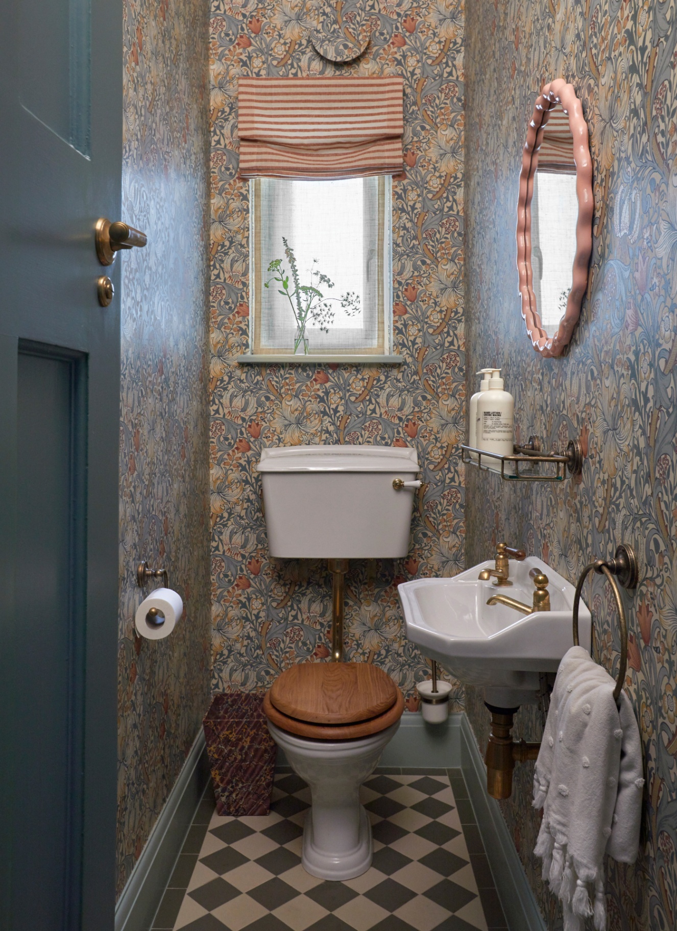 a downstairs toilet with pale blue, cream and orange william morris wallpaper, a small window with apale pink and white stripe blind, a white toilet with a wooden seat, blakc and white checkerboard floor tiles and a pale pink wavy round mirror