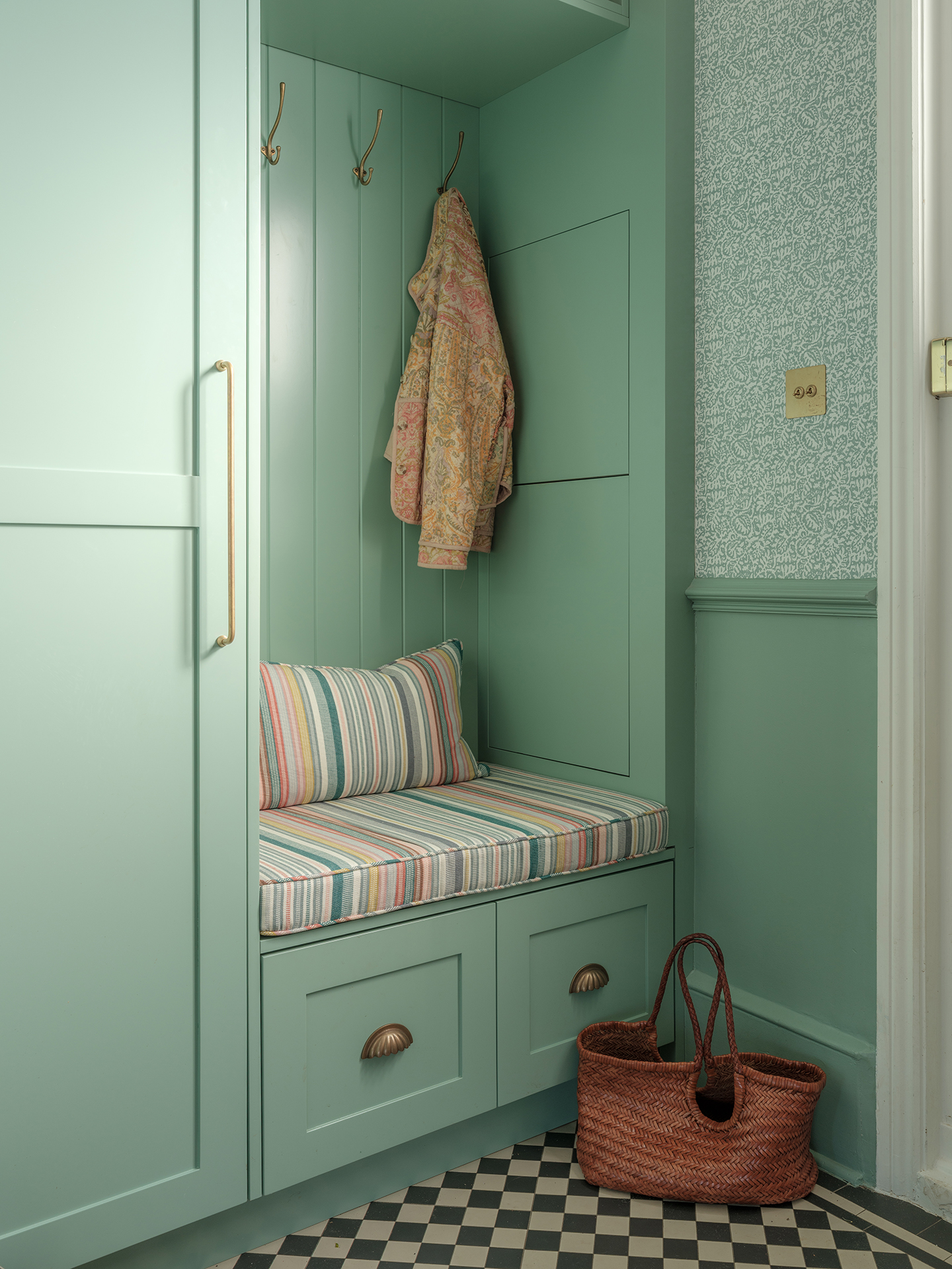 A hallway, with the lower half of walls painted green and the upper half in green wallpaper, with a smal built-in bench seat with a striped cushion. A coat hangs up and a bag is on the tiled floor