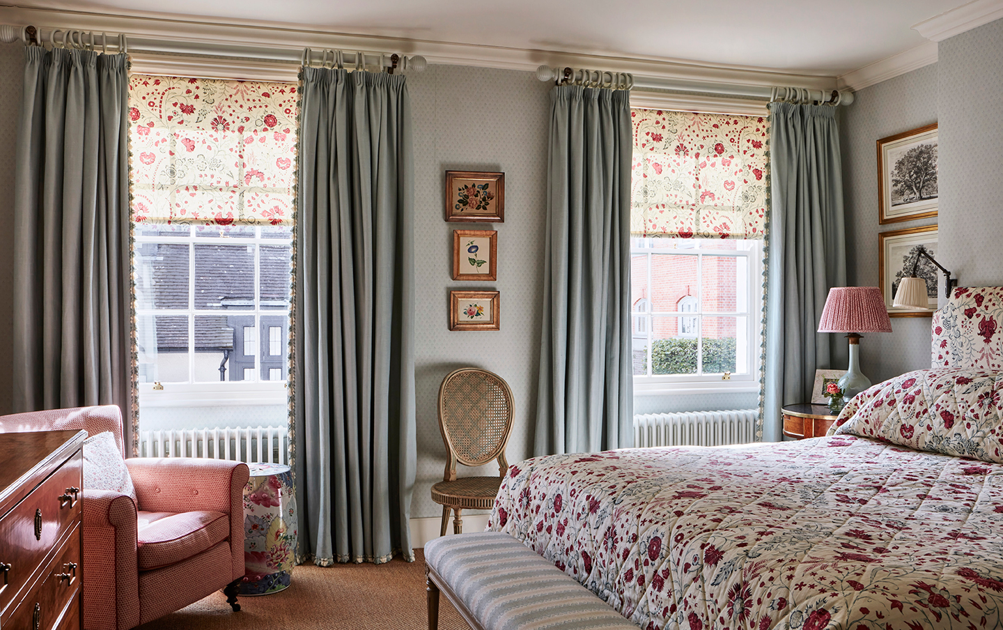 A large blue bedroom with a double bed and two sash windows. The walls and curtains are blue while the blinds, bedspread and headboard are in a red floral chintz fabric. There is a striped bench at the end of the bed and an armchair in the corner