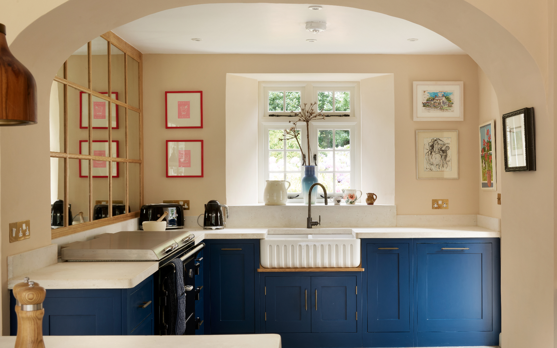 a small kitchen with dark blue cabinetry, cream walls, a belfast sink and a wooden paned mirror on the left wall