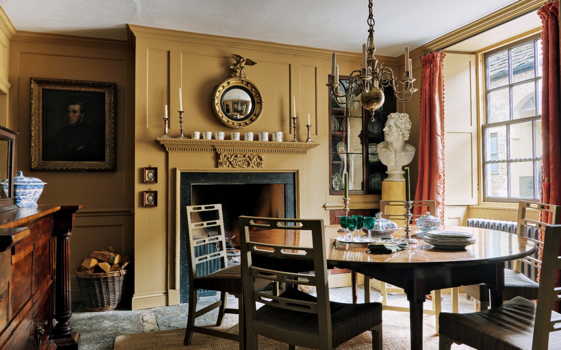 Period dining room featuring mustard yellow panelled walls, a marble-lined fireplace, a round dining table, red window drapes, a classic bust statue, and a brass chandelier.