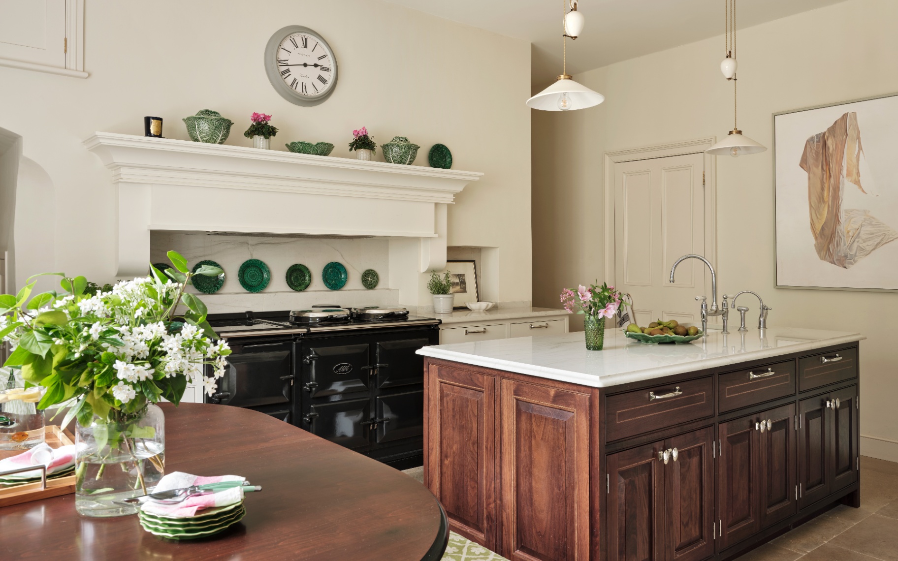 a country kitchen with dark wood cabinetry, a kitchen island, a black aga, green plates on the wall, a table with a vase of flowers and white pendants