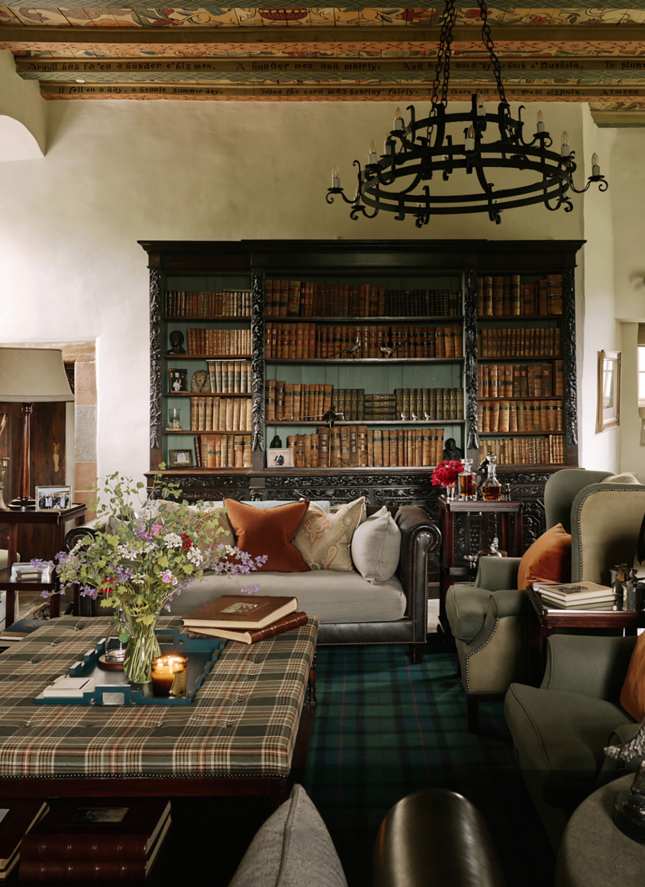 A cozy, traditional room featuring a dark wood bookcase filled with antique books, a grey sofa with orange cushions, green armchairs, a plaid ottoman coffee table, a wrought-iron chandelier, and an ornate painted ceiling