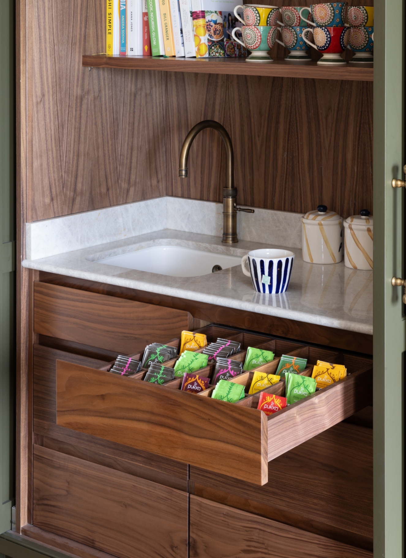 Bespoke walnut kitchen drawer with custom dividers filled with colourful tea bags, situated below a marble countertop prep sink with brass tapware and cookbook shelving