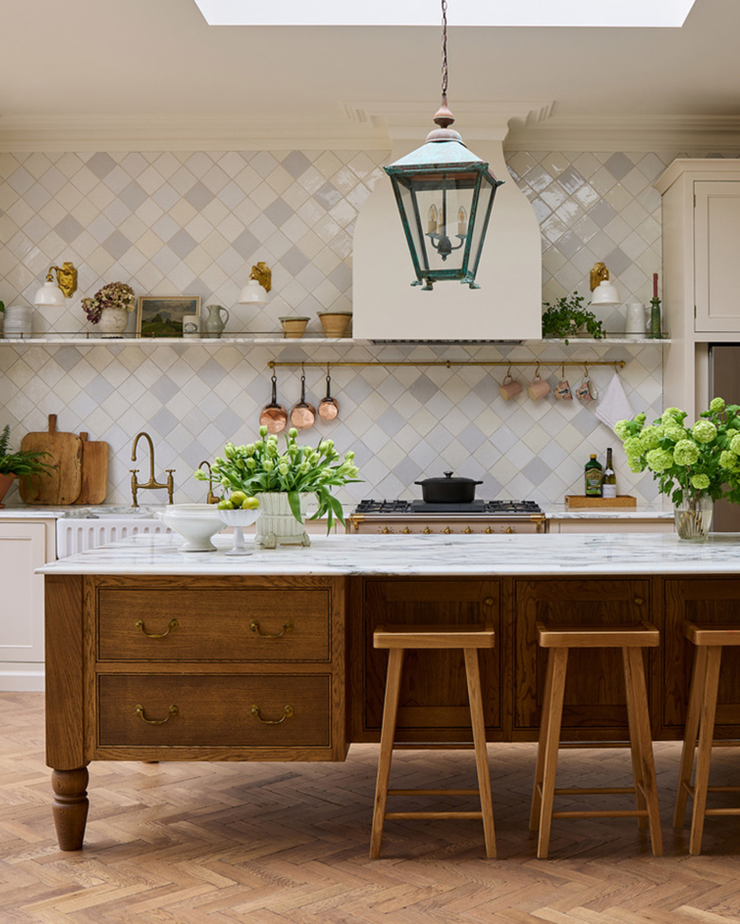 a neutral kitchen with parquet flooring, a large wooden kitchen island with 3 wooden stools, a large blue lantern style pendant, diamond shape white tiled splashback and hanging copper saucepans