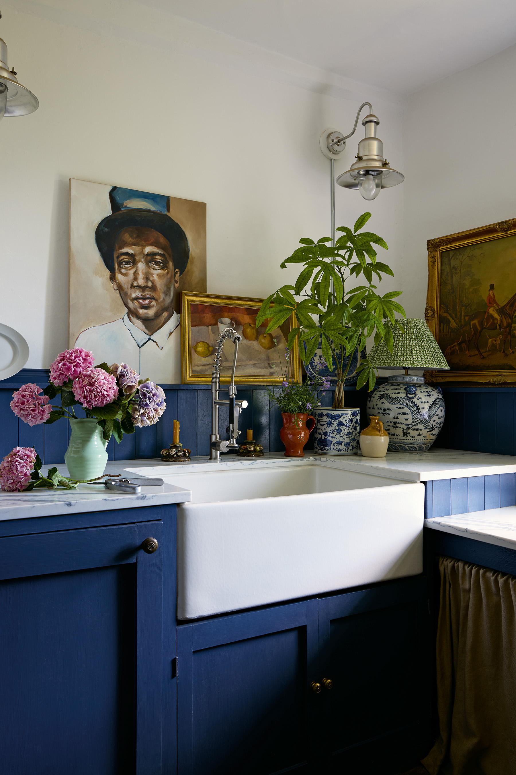 a close up of a utility room area with a ceramic butler sink set in dark blue kitchen cabinets, above are plants and flowers in pots and jugs, a large portrait painting and still life painting and a industrial-style wall lamp