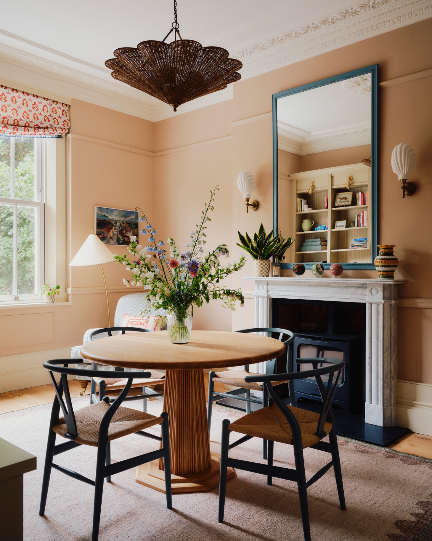 a dining room with a round wooden dining table and 4 dining chairs, a large rattan light hanging above it, a white fireplace with a large blue mirror above it and two shell shape sconces either side