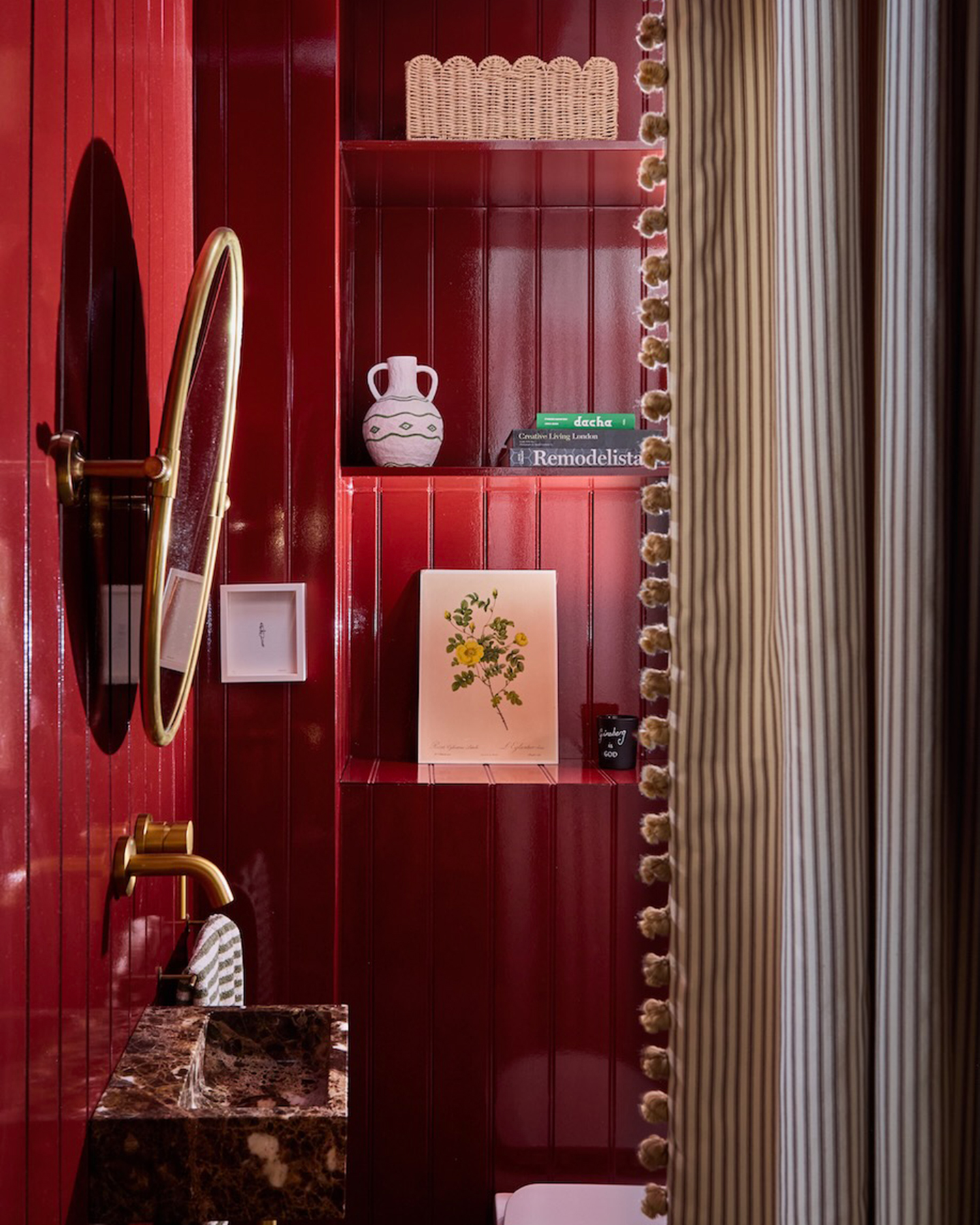 a small, narrow bathroom with bright red panelled walls, a brown marble sink with a gold circle mirror above it and open shelves with books and a print of yellow flowers