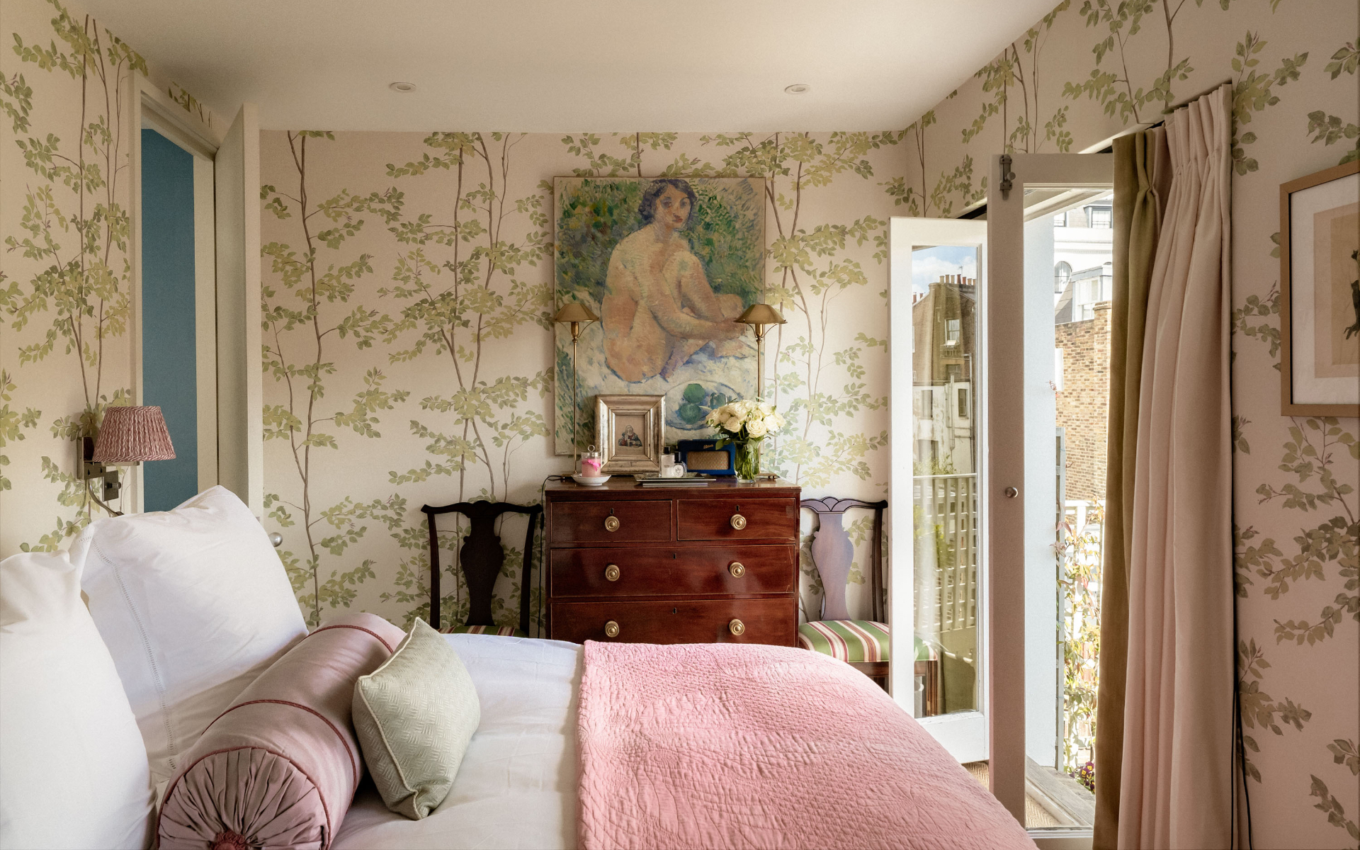 a bedroom with green botanical wallpaper, two large windows wide open onto a balcony, a bed with pink bedding and a paitning above an antique chest of drawers