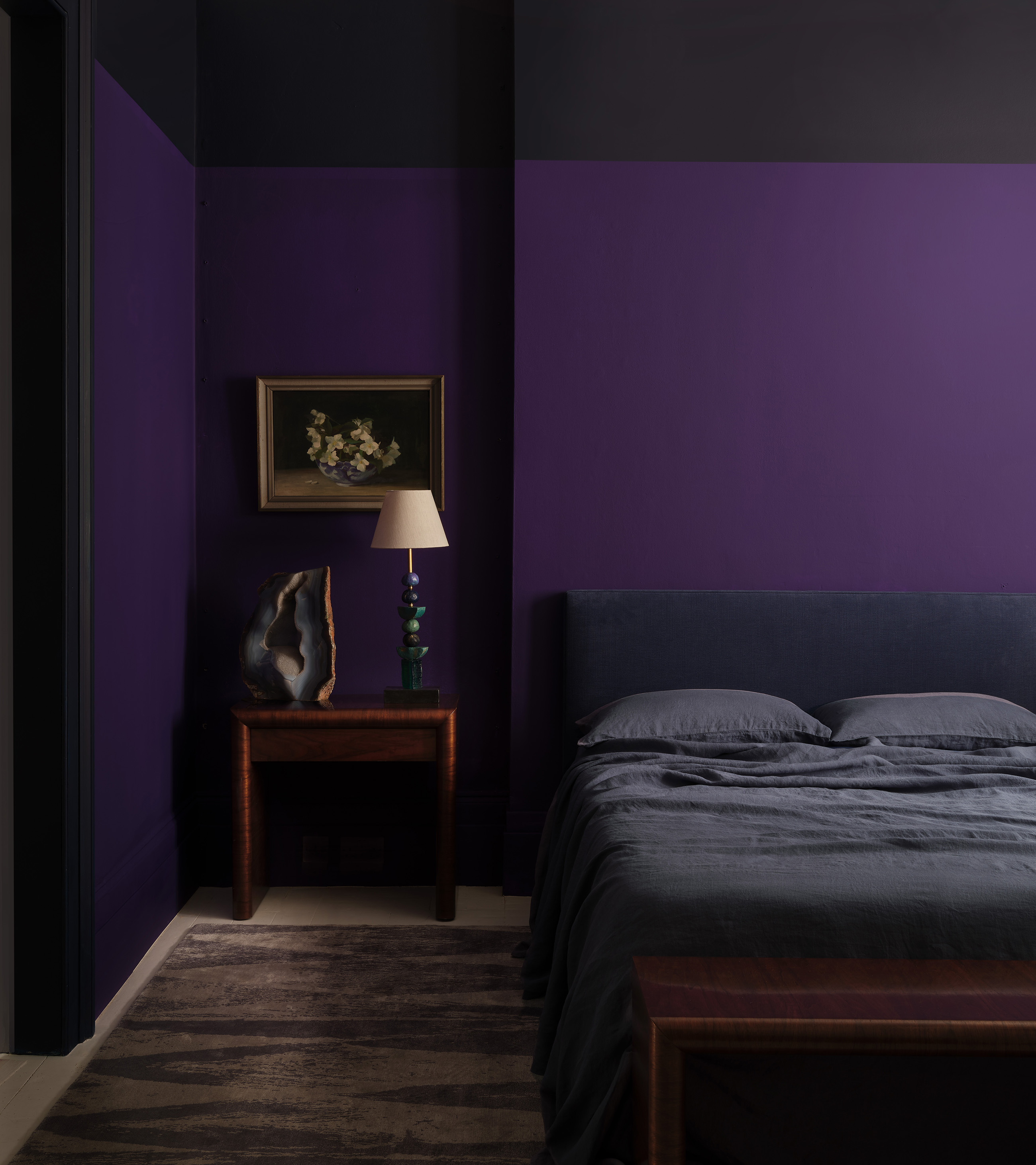 a dark bedroom with black and purple painted walls, a bedside table with a lamp and frame hung above the wall and a double bed with black bedding