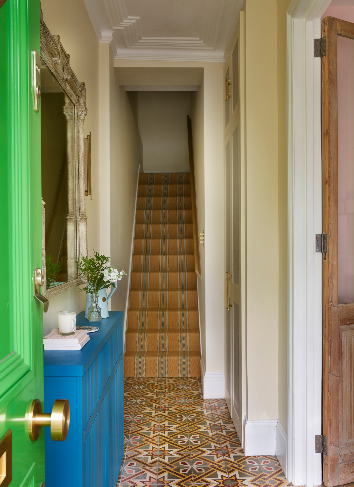 a hallway with a bright green open front door, a blue console table with a candle and vase on top, a mirror above it and stairs with ochre striped carpets