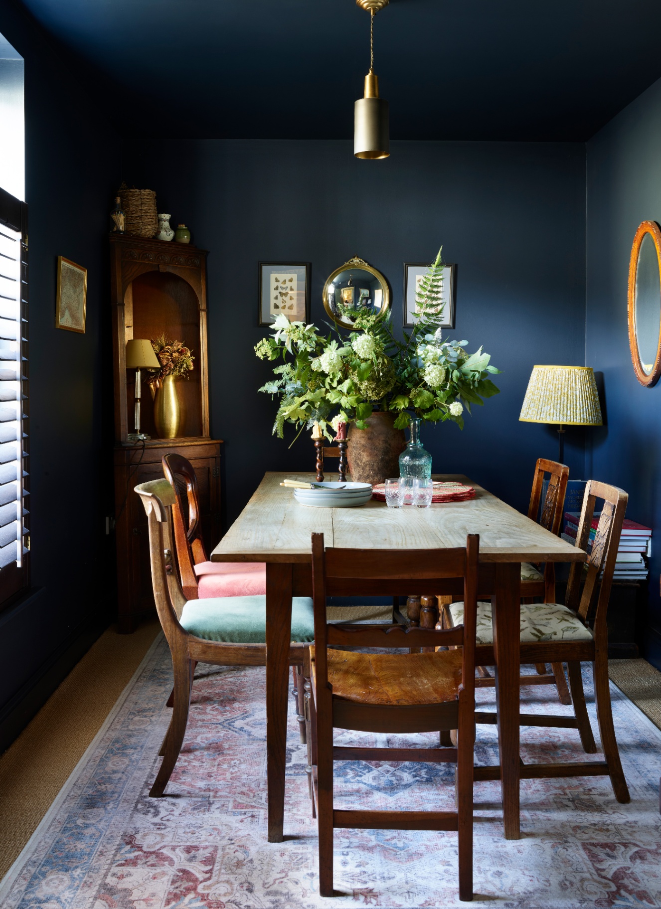 Intimate dining room with dark navy blue color-drenched walls and ceiling, rustic wooden table, antique corner cabinet, and patterned area rug