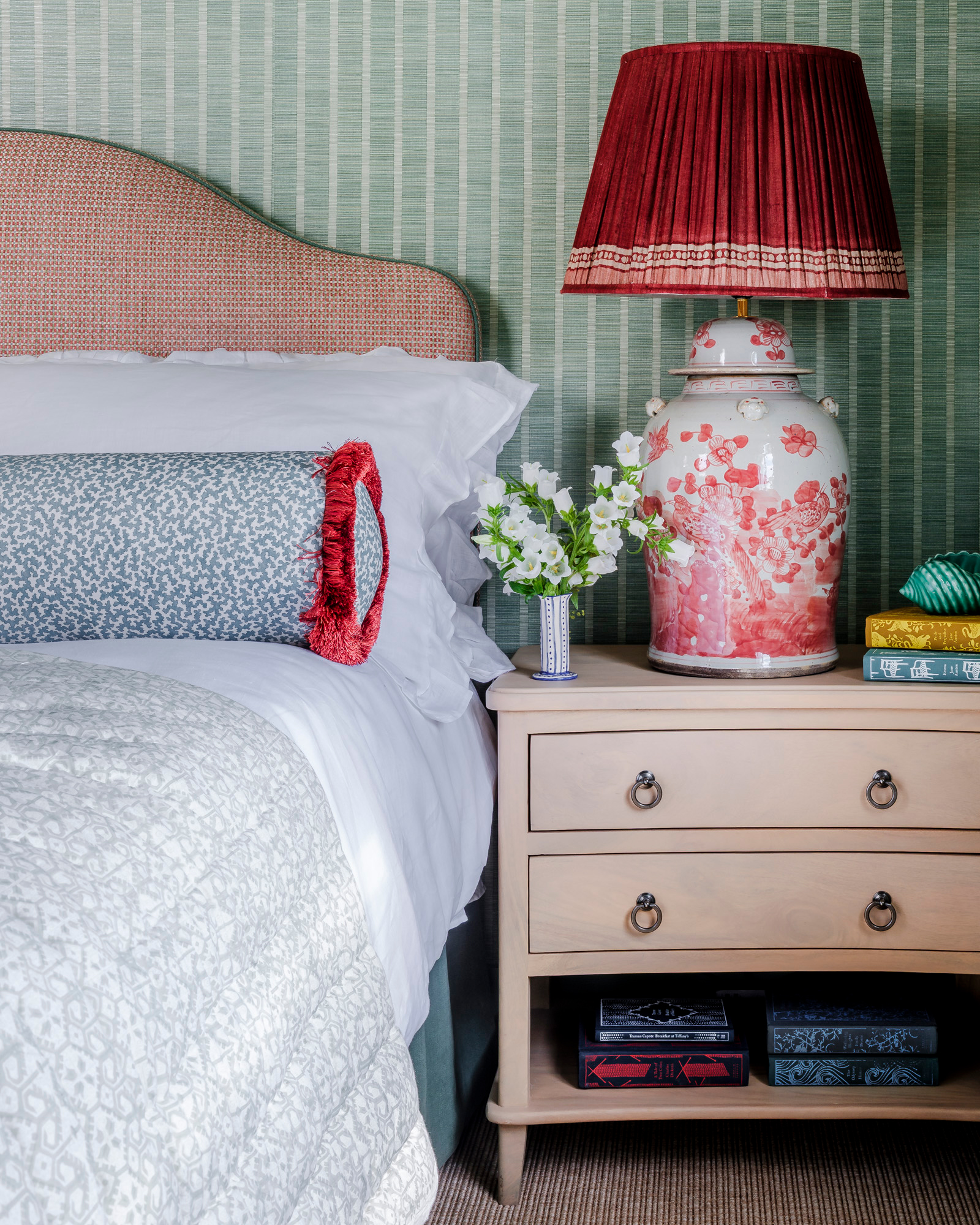 a bedroom with green and white striped wallpaper, a pale wooden bedside table with a large ceramic red and white lamp and a bed with white bedding and a blue and red bolster cushion