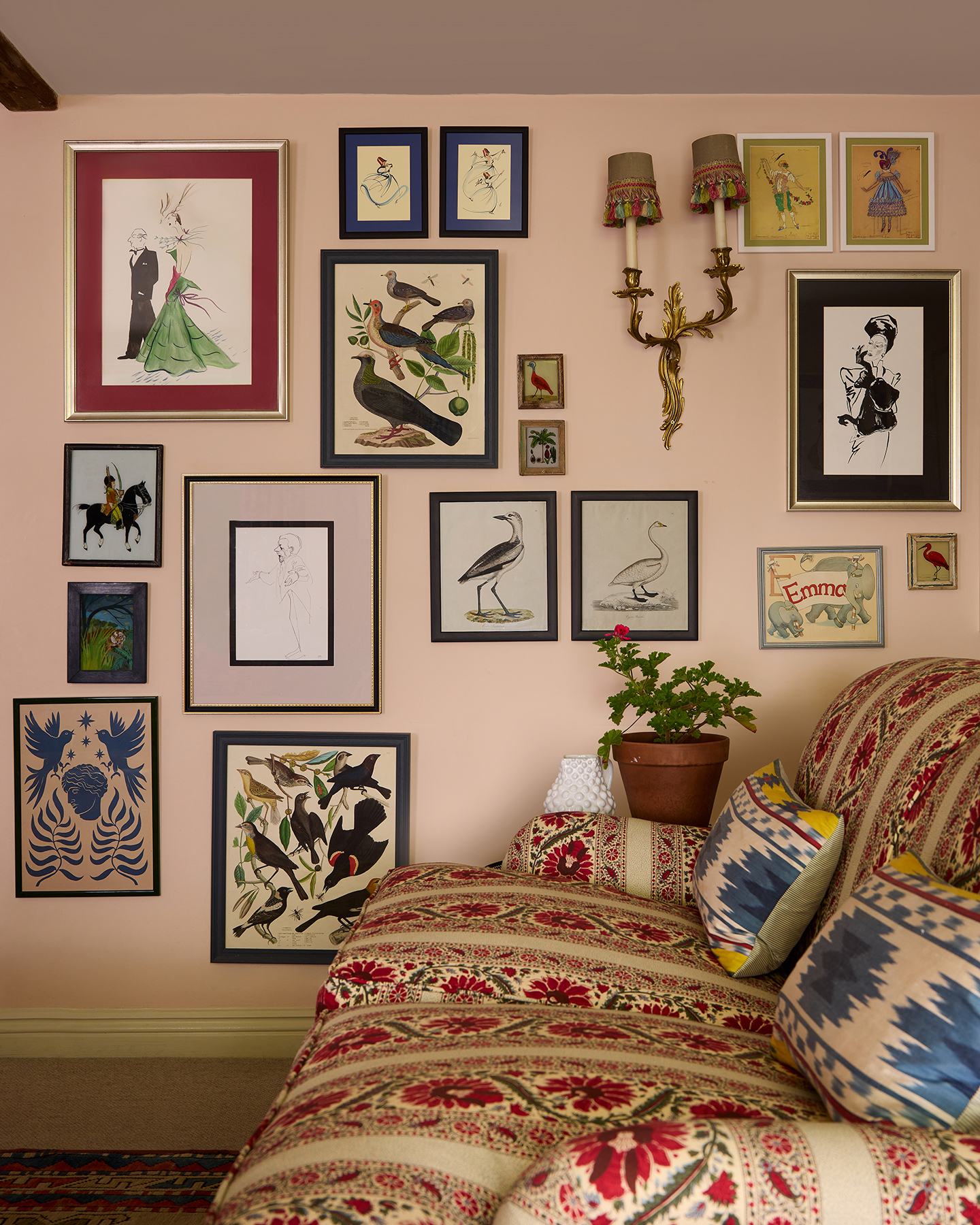 A gallery wall created with a mix of artworks in all shapes and sizes, including illustrations of people, birds, and other animals. An ornate wall light hangs near the centre and in the foreground is a patterned sofa  