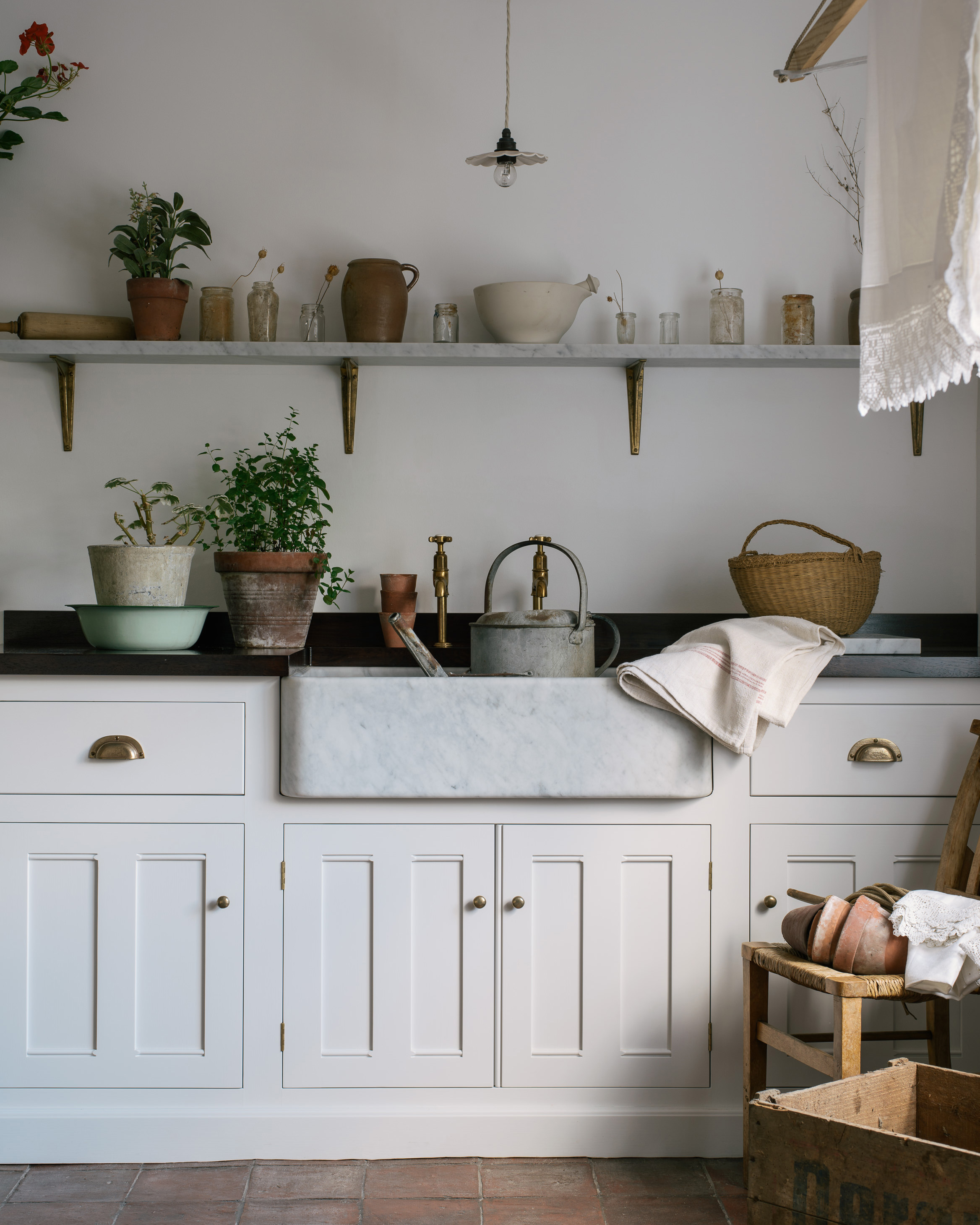 a white utility room with brick floor, a marble belfast sink with a silver metal watering can inside and an open shelf above with lots of jars and pots