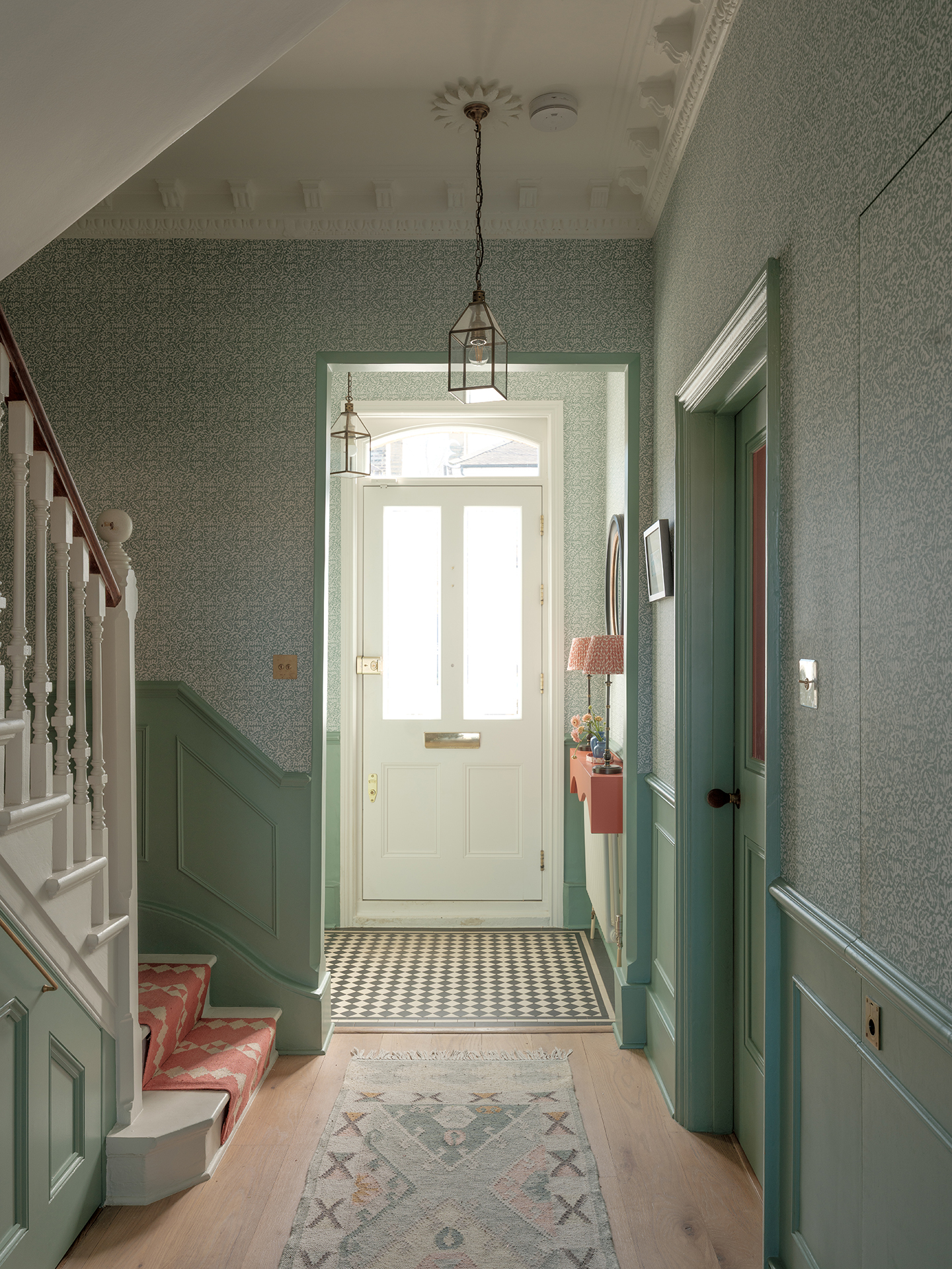 A hallway with the lower half of walls painted green and the upper half in green wallpaper. Stairs start on the left and a runner in the foreground leads to a tiled entryway and the front door. A lantern pendant hangs above 