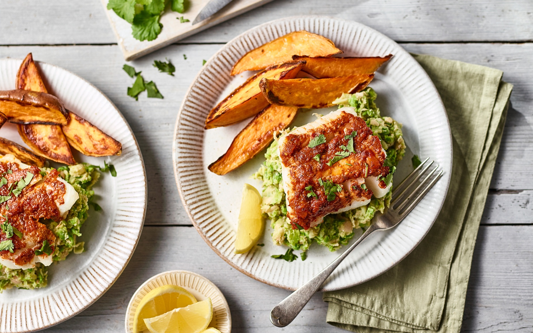 a white dinner plate with cod, pea mash and sweet potato wedges with a silver fork and a sage green napkin