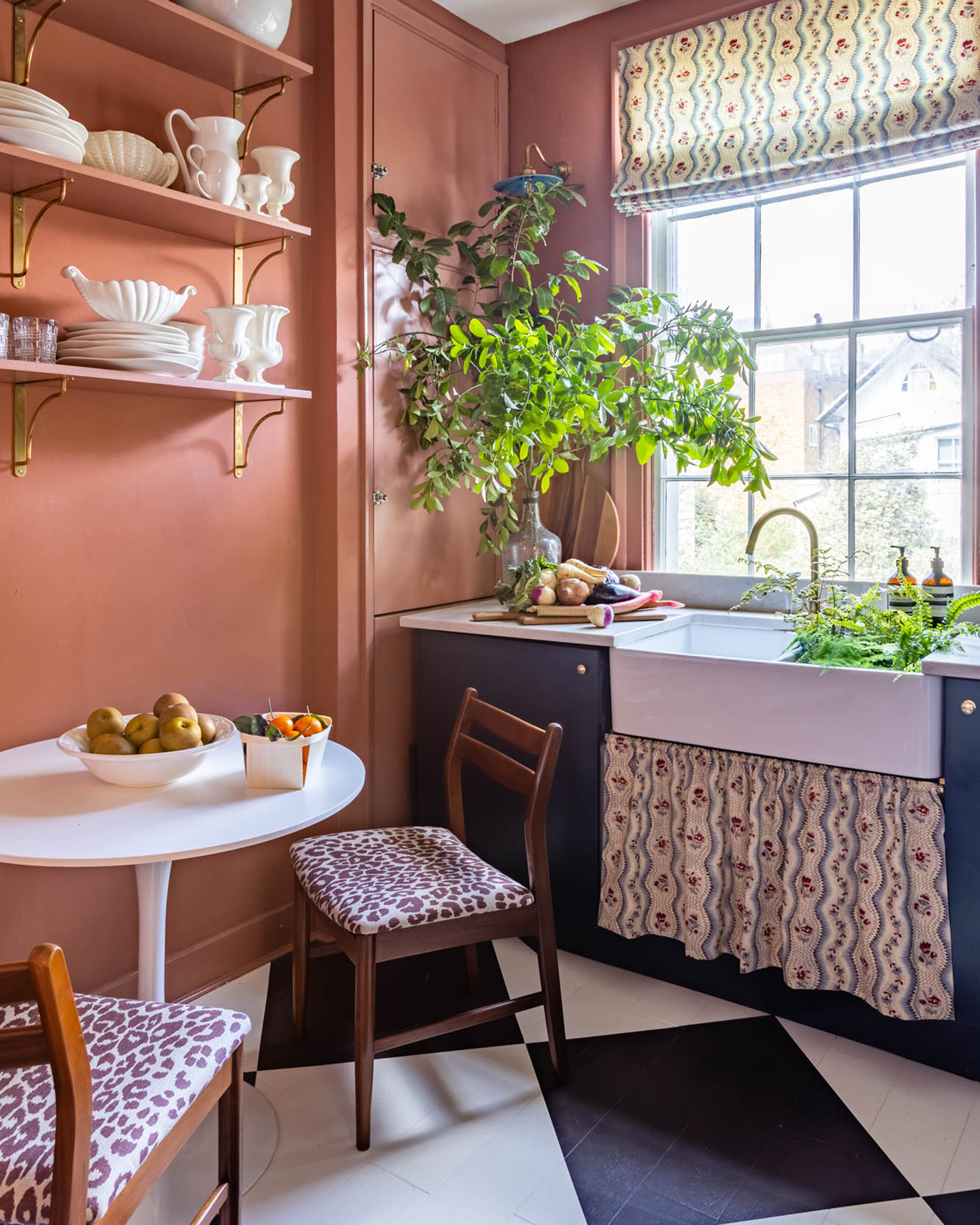 a kitchen with black and white checkerboard floors, a small white round table with wood and leopard print dining tables and a belfast sink with a patterned fabric skirt and matching blinds
