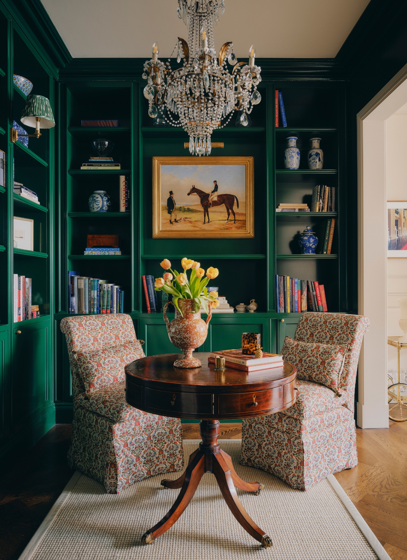 A formal dark green library corner featuring floor-to-ceiling green bookshelves, a central mahogany pedestal table, two upholstered chairs, and a crystal chandelier