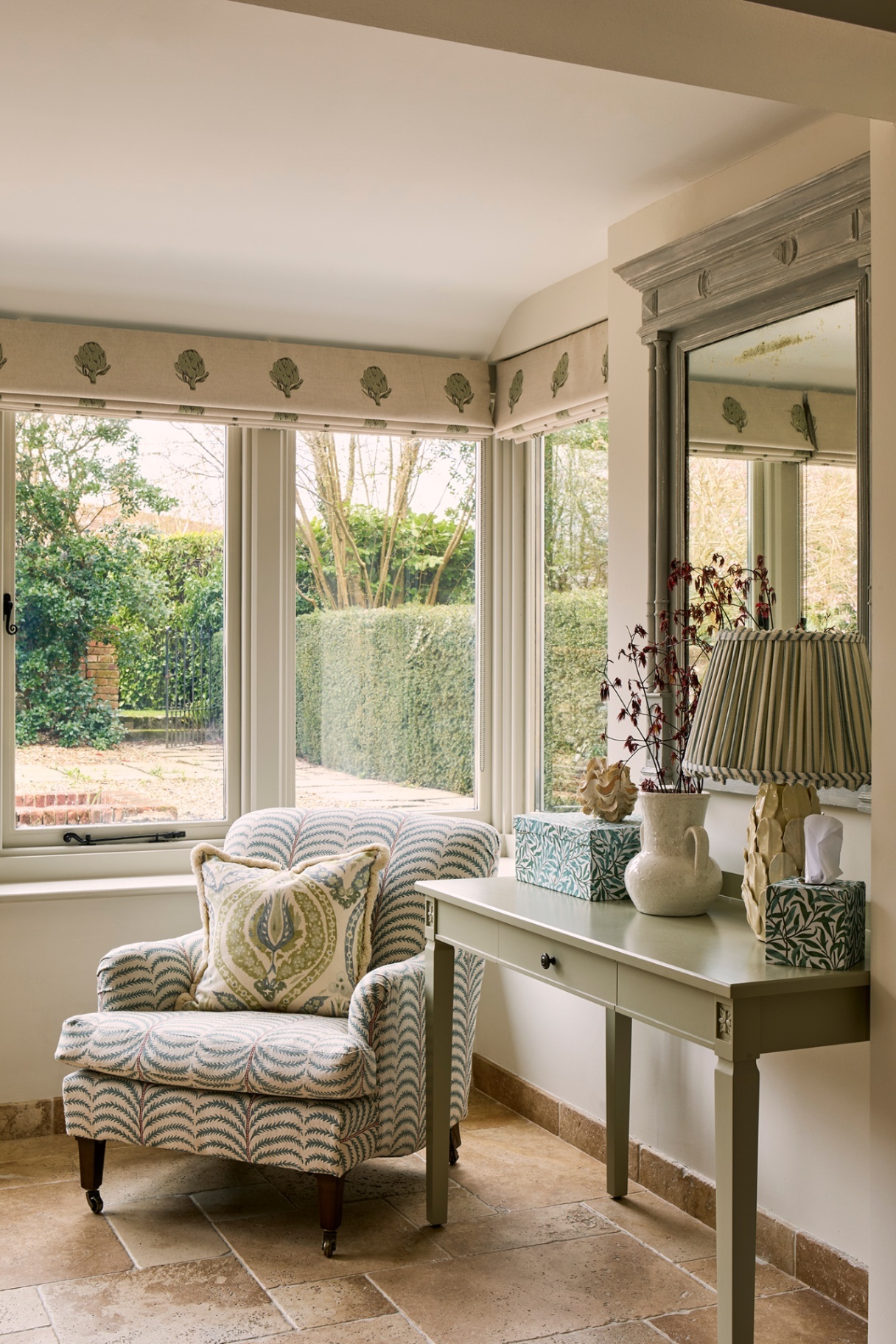 A blue and white print chair with a fringed cushion is set in a corner with large windows dressed with blinds made in an artichoke-print fabric. To the right is a console table topped with a vase, lamp and other accessories, and above hangs a mirror