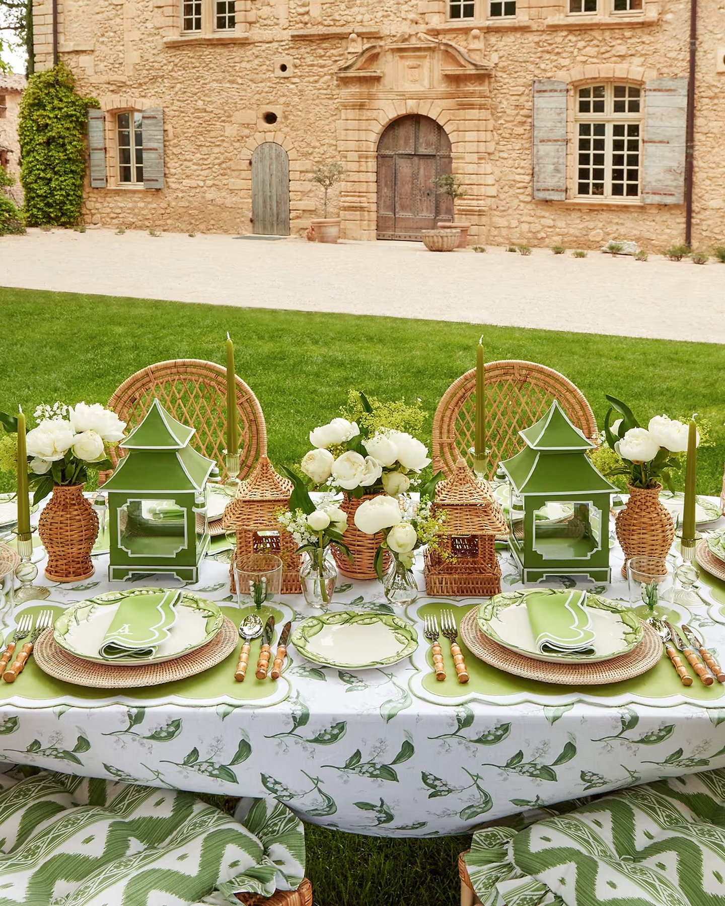 an outdoor dining table with a white and green tablecloth, green placemets with rattan charger plater, rattan lanterns and vases filled with white flowers and two large green lanterns