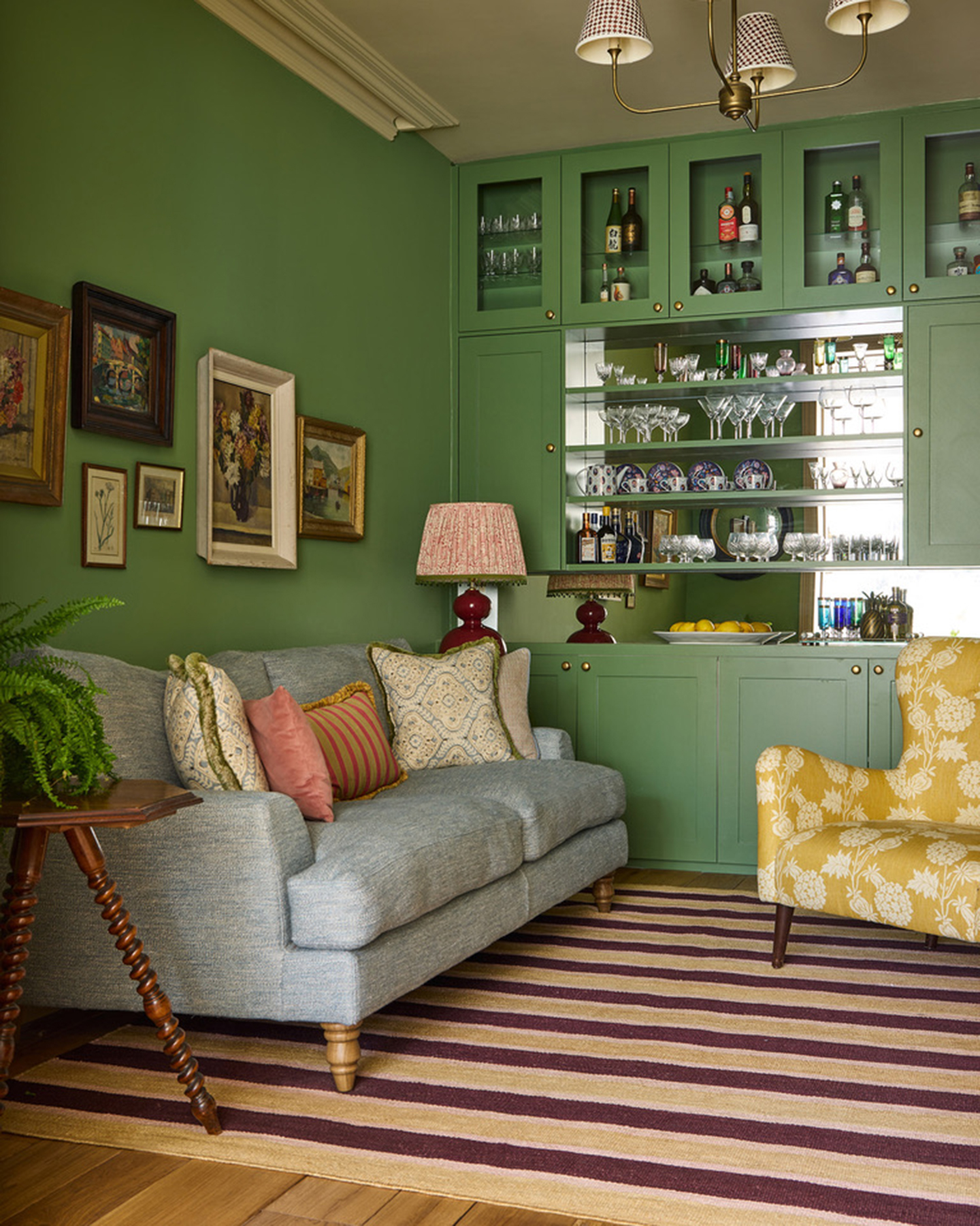 A LIVING ROOM WITH BRIGHT GREEN WALLS, a yellow and purple stripe rug, a pale blue sofa and build in shelving with glasses and bottles