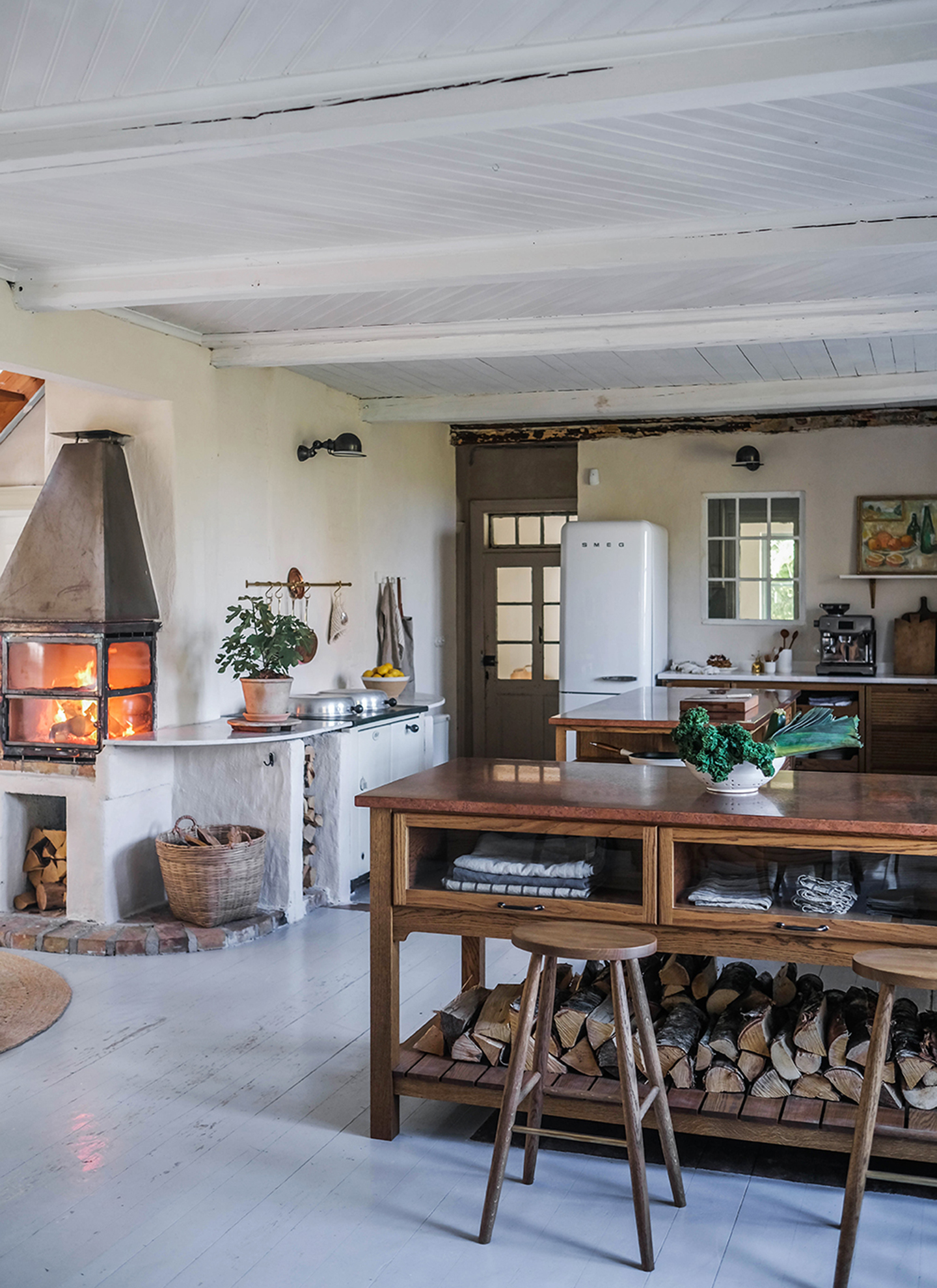 a kitchen with a wooden kitchen island with logs stored under it, white wooden floors and a log burner