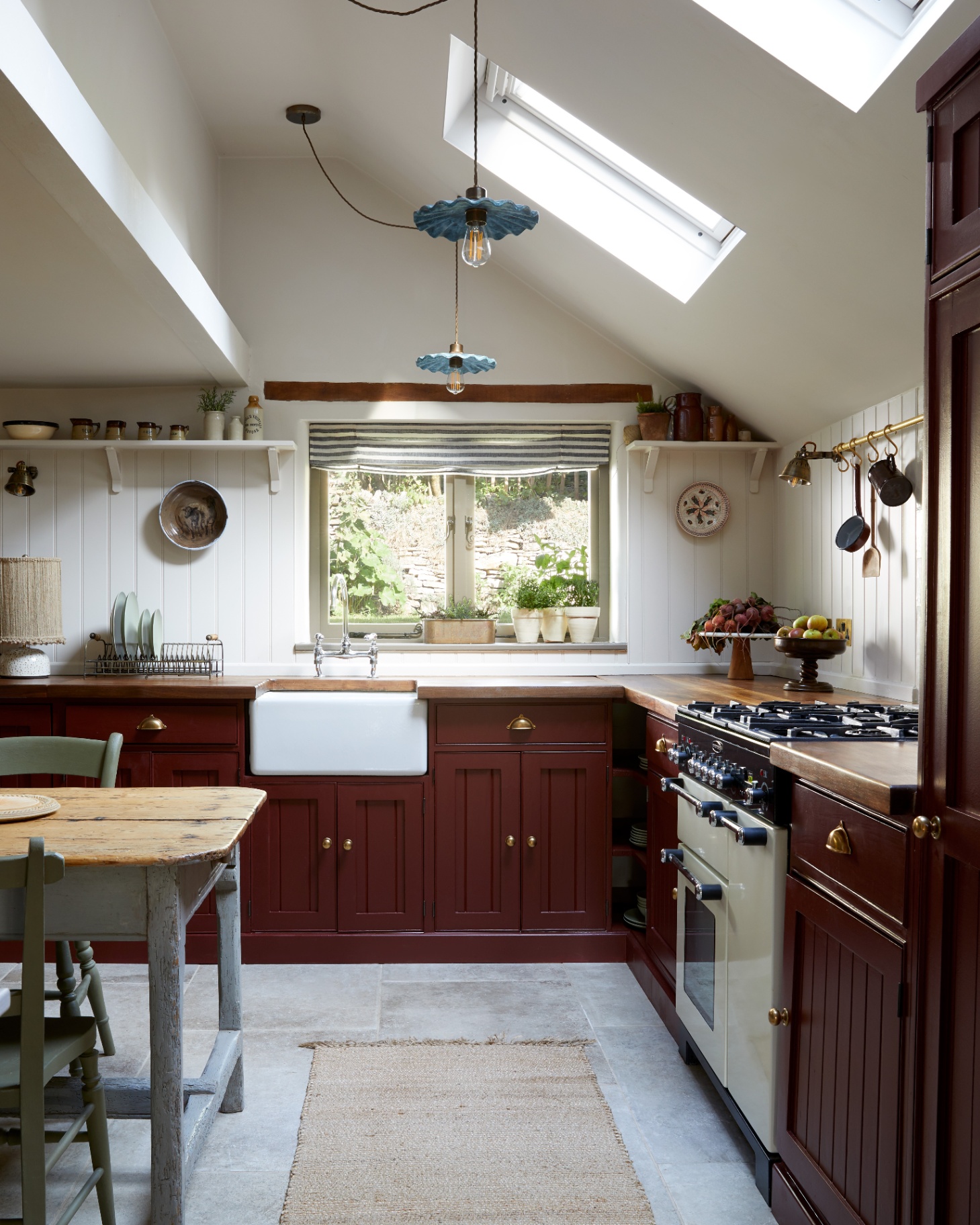 a kitchen with burgundy painted cabinets, a jute runner rug, two pendant lights with blue frilled shades and a farmhouse kitchen table with grey chairs