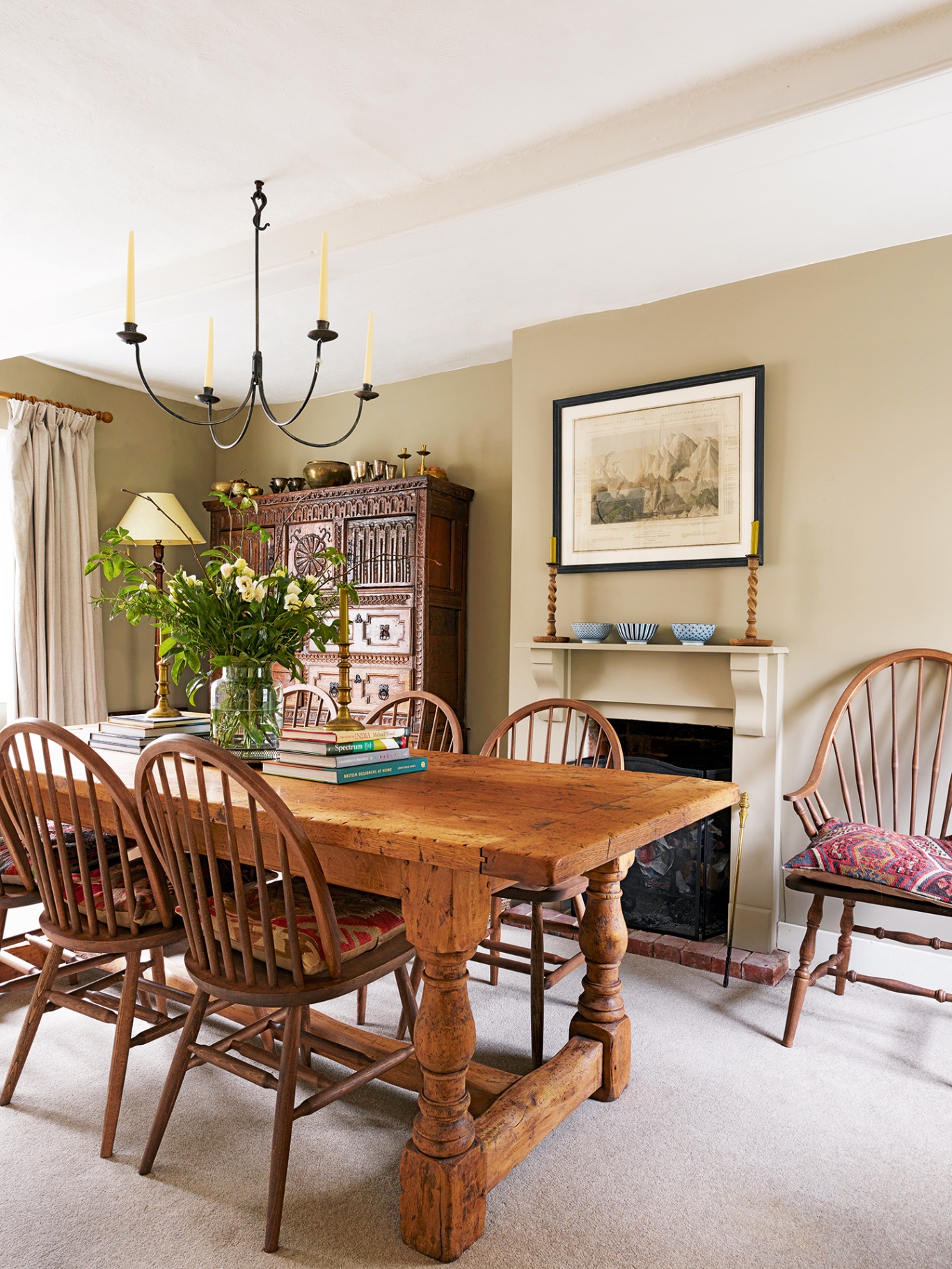 A neutral dining room with a large dining table and six spindleback chairs in the foreground, and a large antique chest, fireplace and wooden carver chair in the background. A simple four-arm candle holder hangs above the table