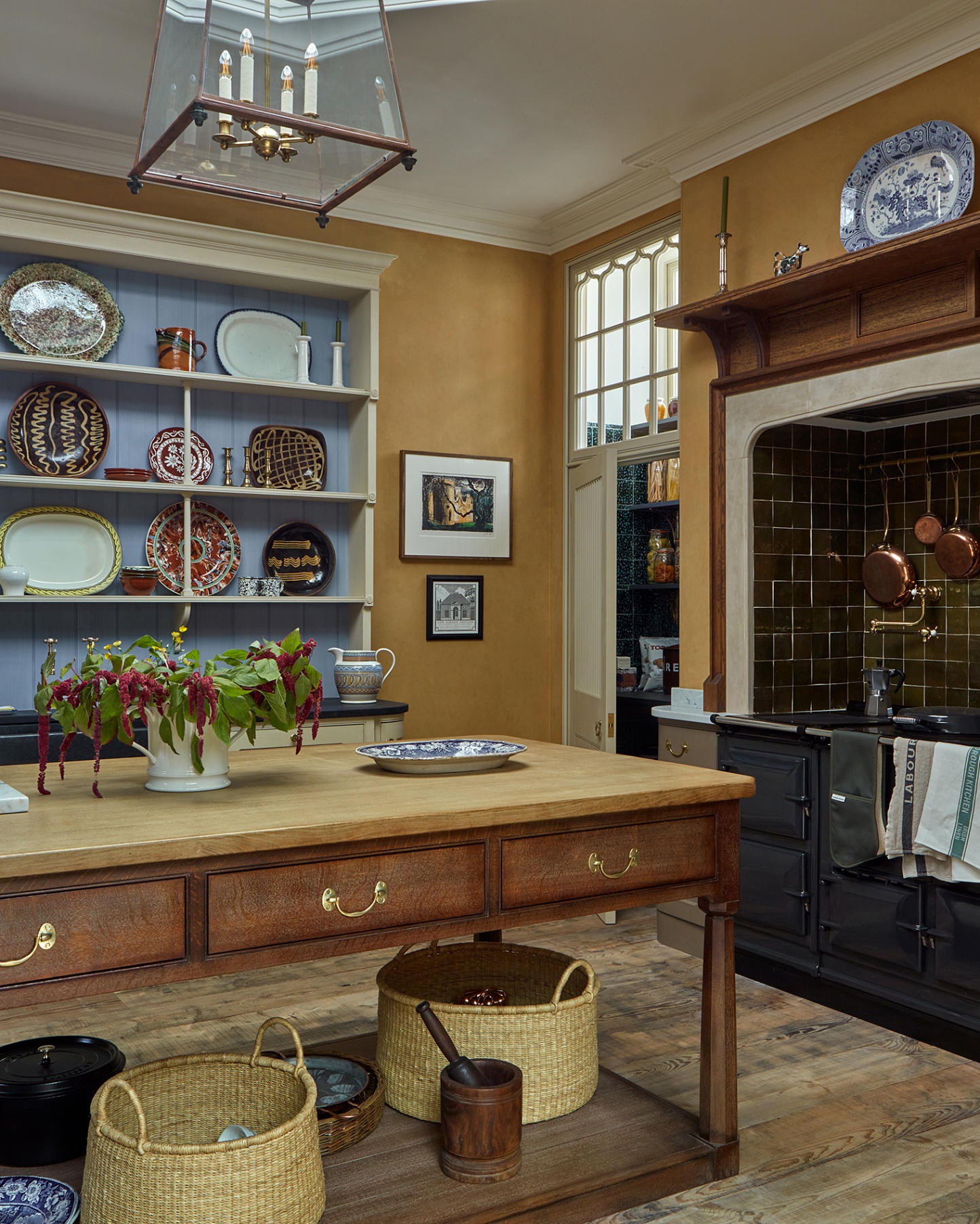 a kitchen with a large dark wooden kitchen island with wicker baskets underneath, a large white and blue painted open dresser on the back wall full of plates and a large navy range cooker with green tiled splashback