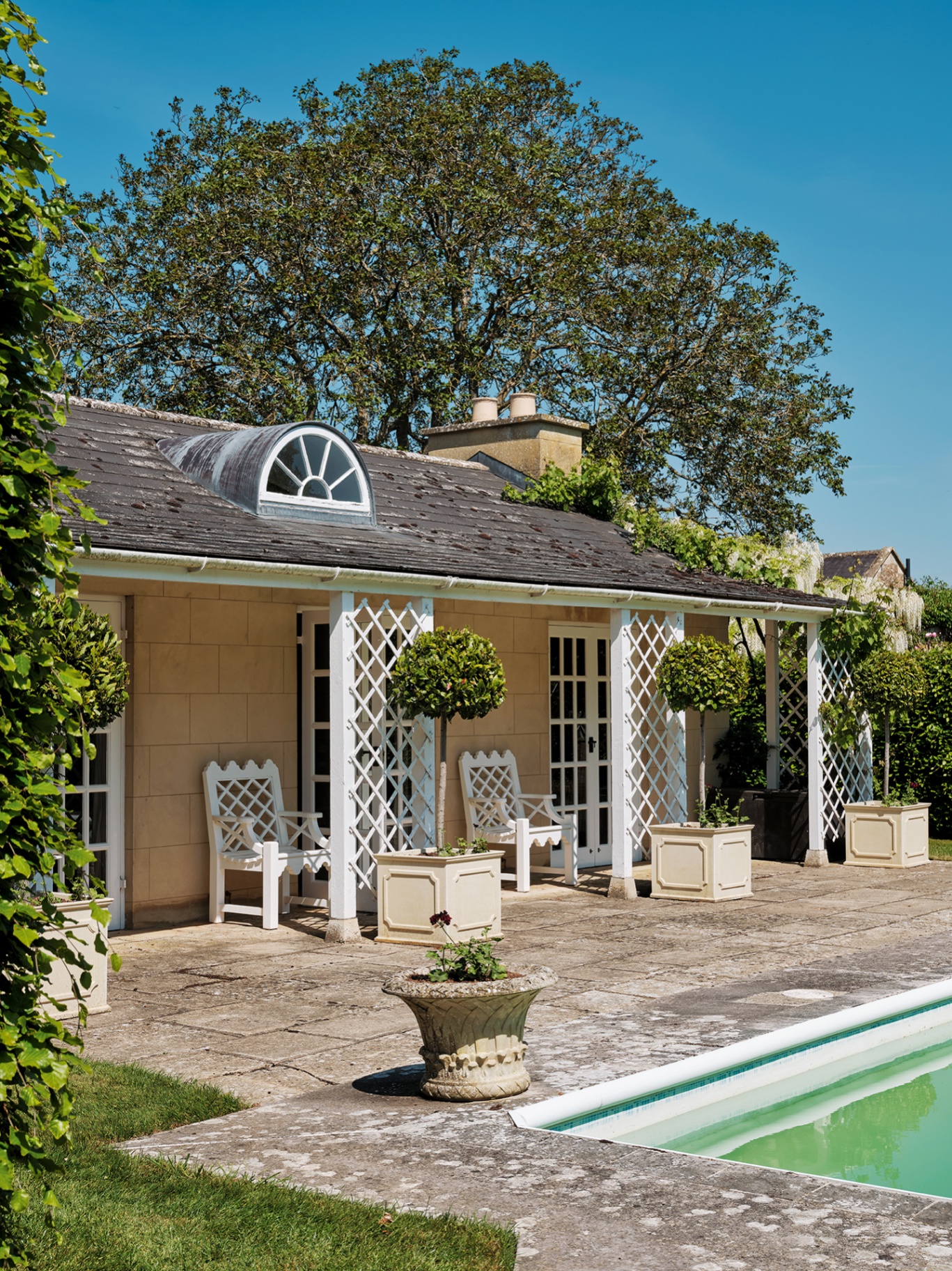 a pool house with a semicircular dormer window and trellis panels and lattice chairs outside. Large square planters line the front with bay trees in them. 