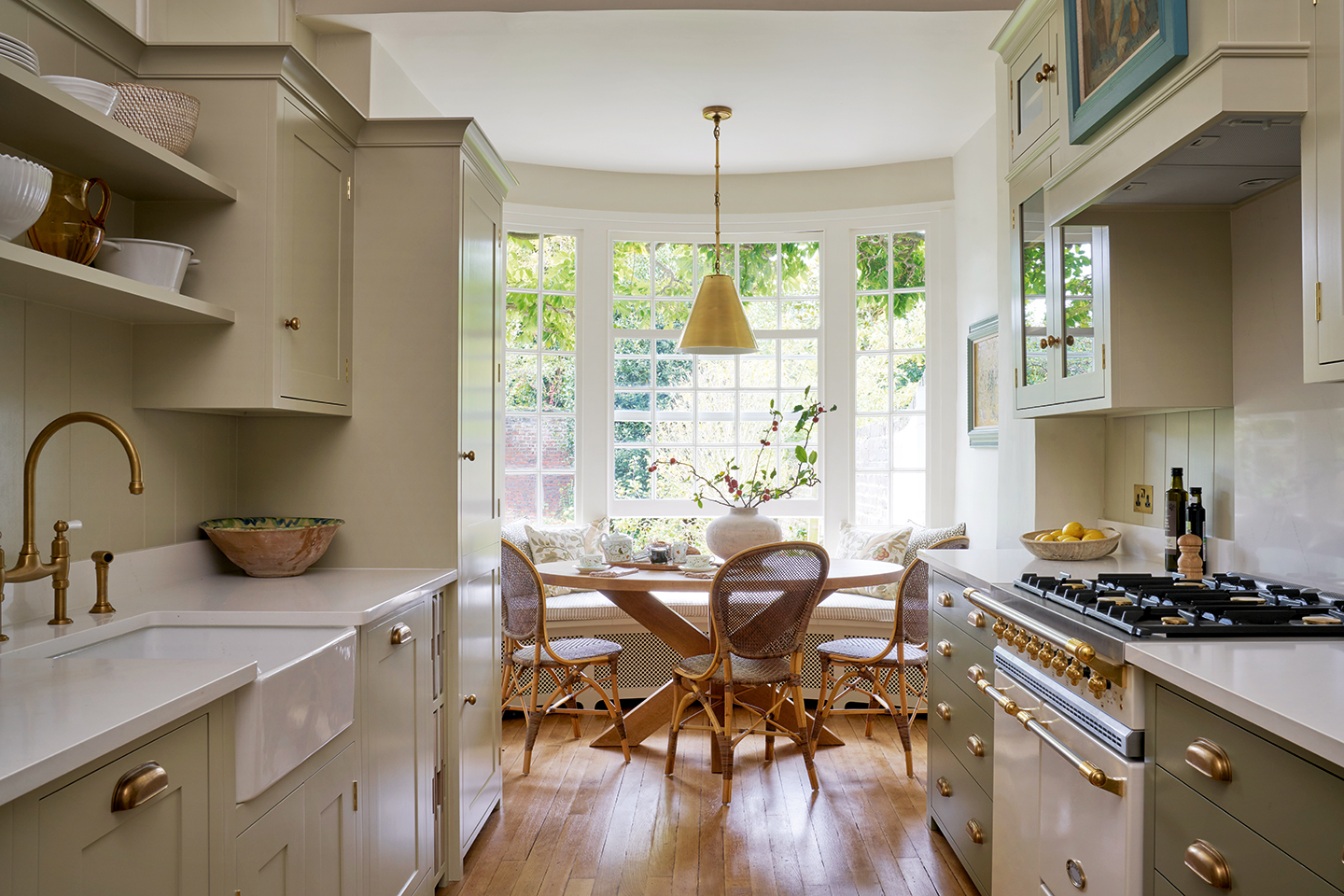 A wide galley kitchen with cabinets and open shelves in the foreground, encompassing a cooker and butler sink, while in the background is a circular table paired with dining chairs and built-in banquette seating in front of large windows