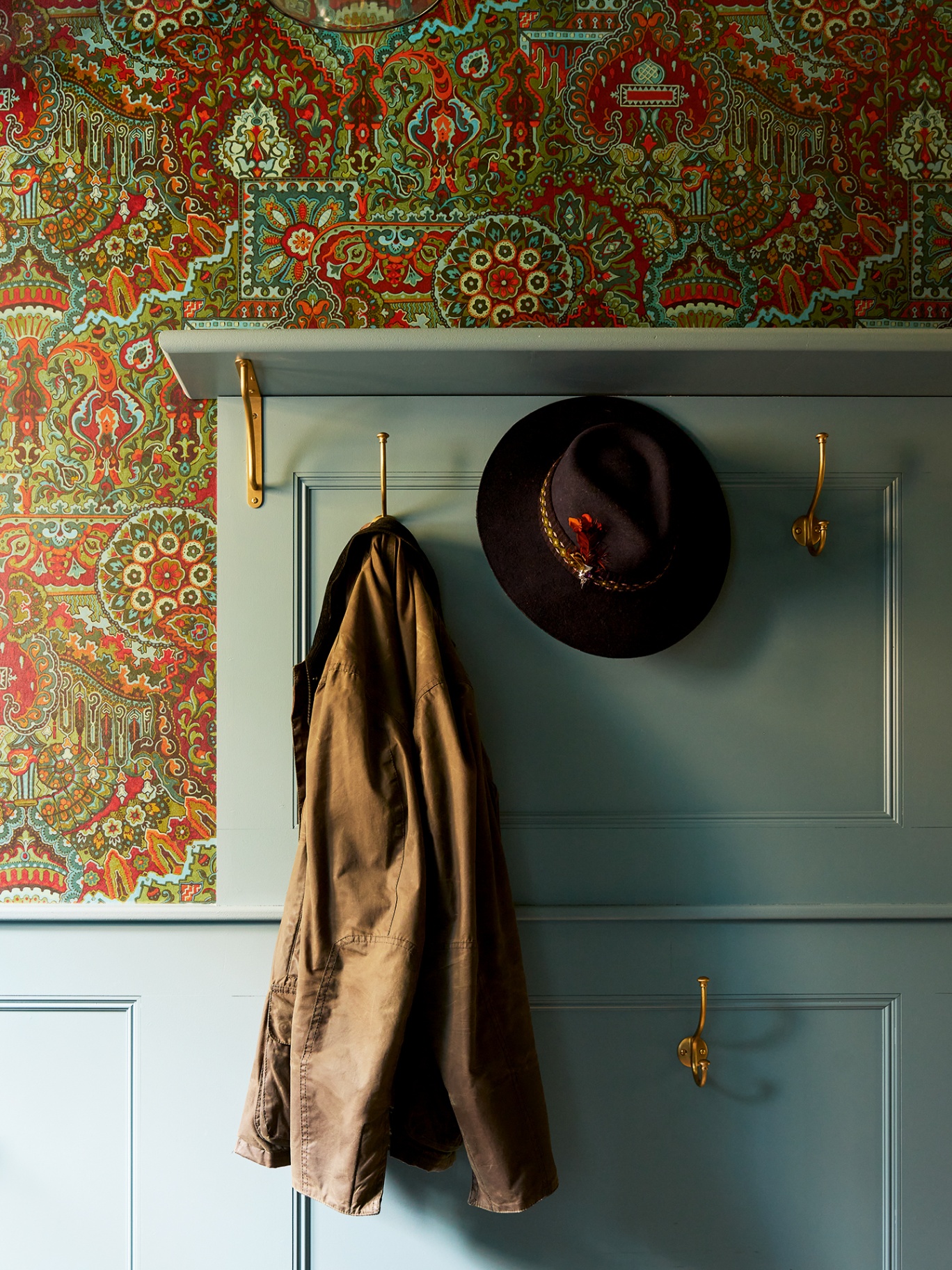 A close up of a cloakroom with patterned green, orange and red wallpaper on which sits light blue painted panelling with gold hooks fixed to it. Hanging up is a brown fedora and a brown wax jacket