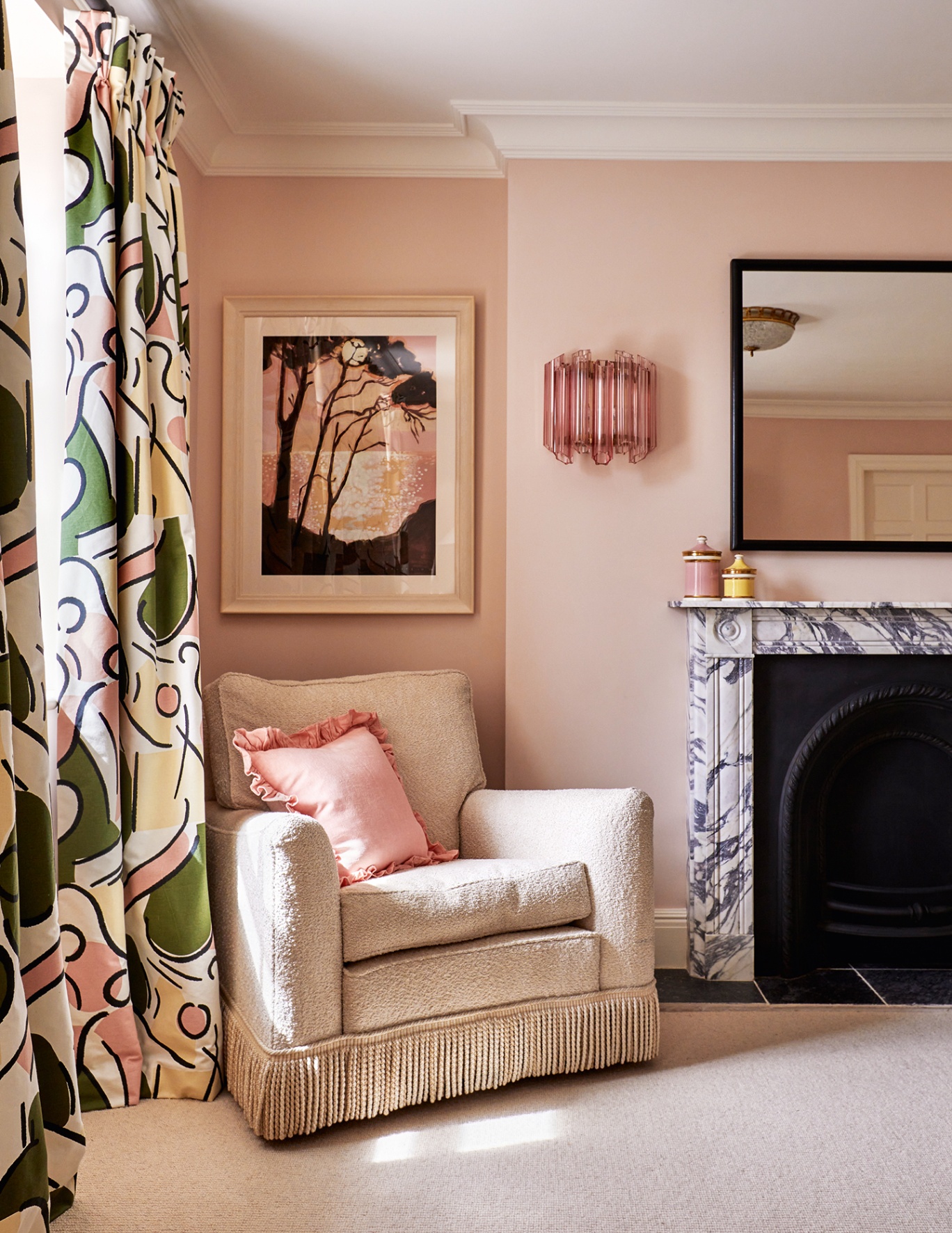 Corner of a pink bedroom with a fringed armchair, marble fireplace and graphic curtains. A landscape print, sculptural pink wall light and rectangular mirror hang on the wall above
