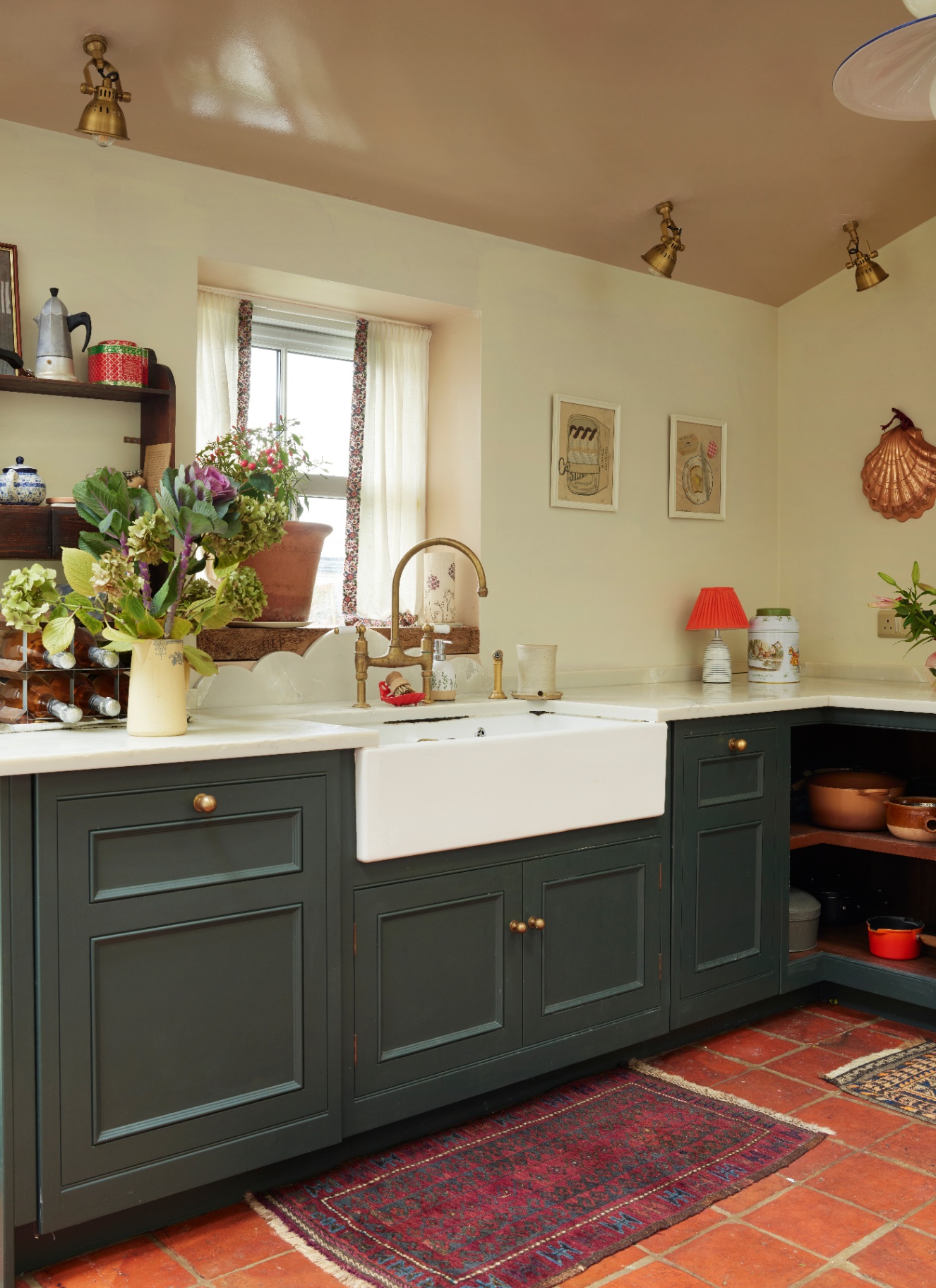 a kitchen with terracotta floor, dark green cabinets, a belfast sink, white walls, gold taks lighting and a brass shell sconce