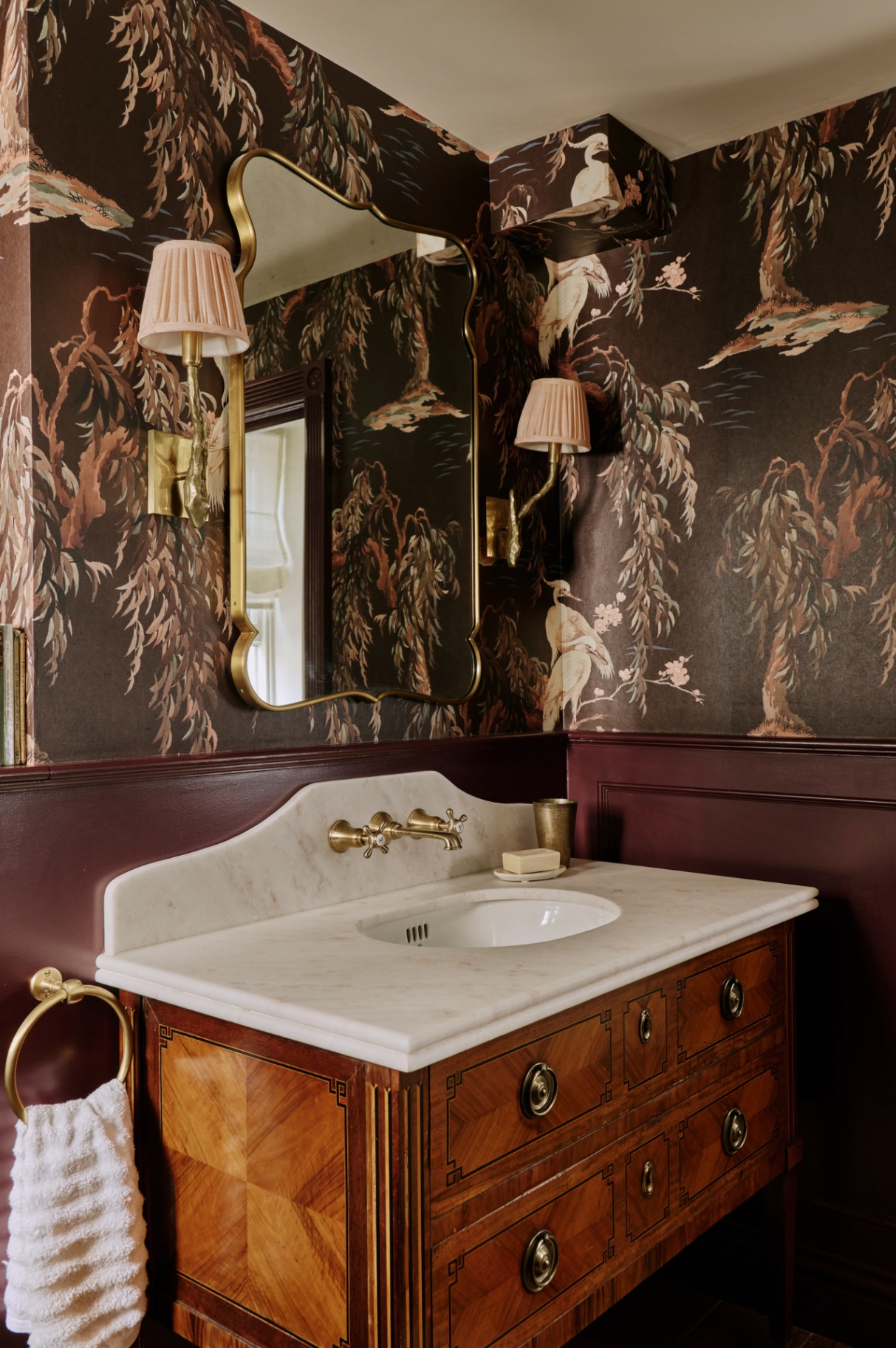 A close up of a cloakroom with a wooden vanity with marble basin top. Above is a gold frame mirror and two wall lights. The walls are covered in a wallpaper with a large-scale tree and bird print