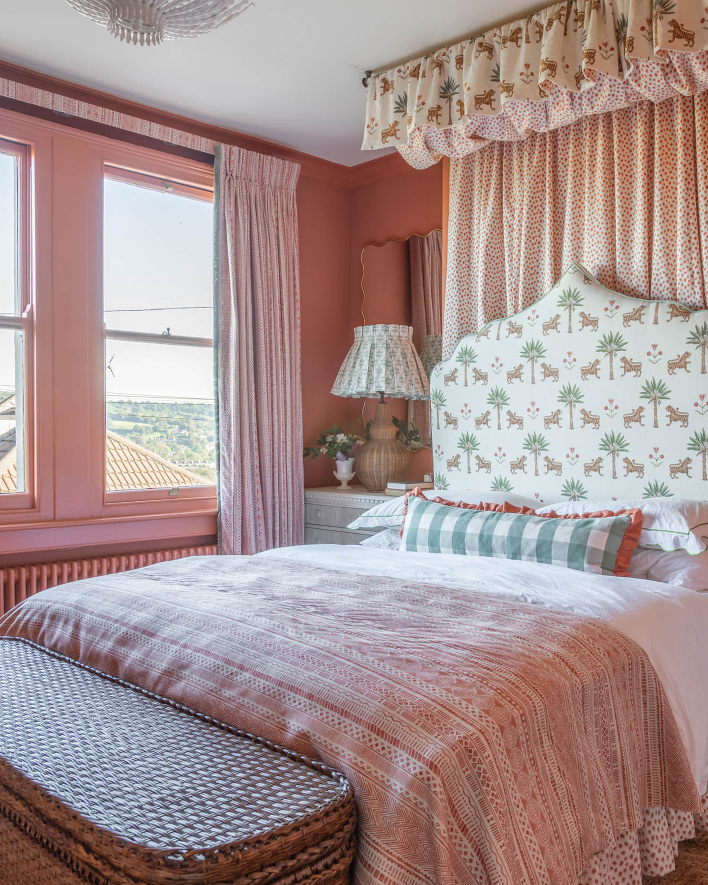 a bedroom with terracotta red walls, a large bed with a white and green patterned headboard, a pink bedspread and a patterned canopy