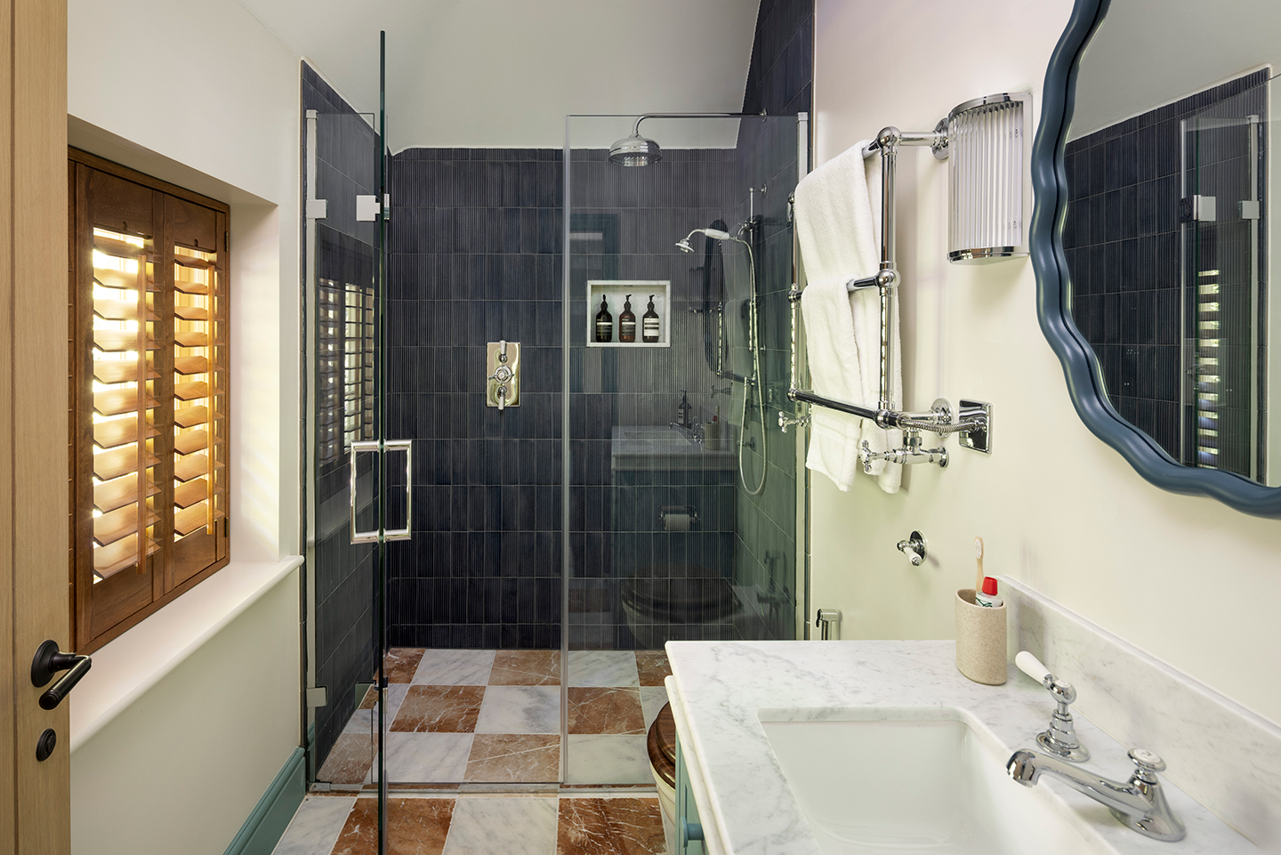 a shower room with brown and white marble checkerboard floor and a glass shower at the back of the room with black wall tiles. In the foreground is a marble basin, wavy circular mirror, towel rail and ribbed glass wall light