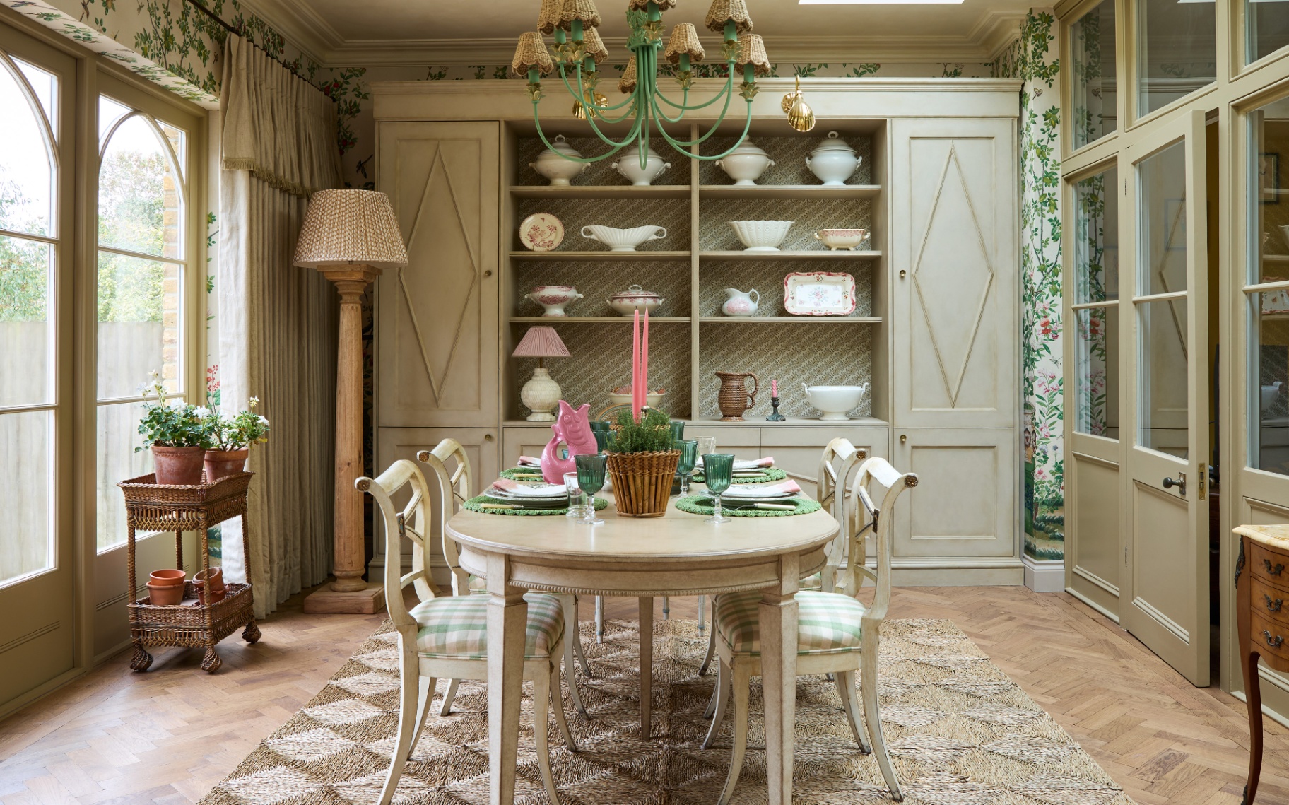 a neutral dining room with a green and rattan chandellier above a pale wood dining table with a pink fish jug and pink candle sticks on top