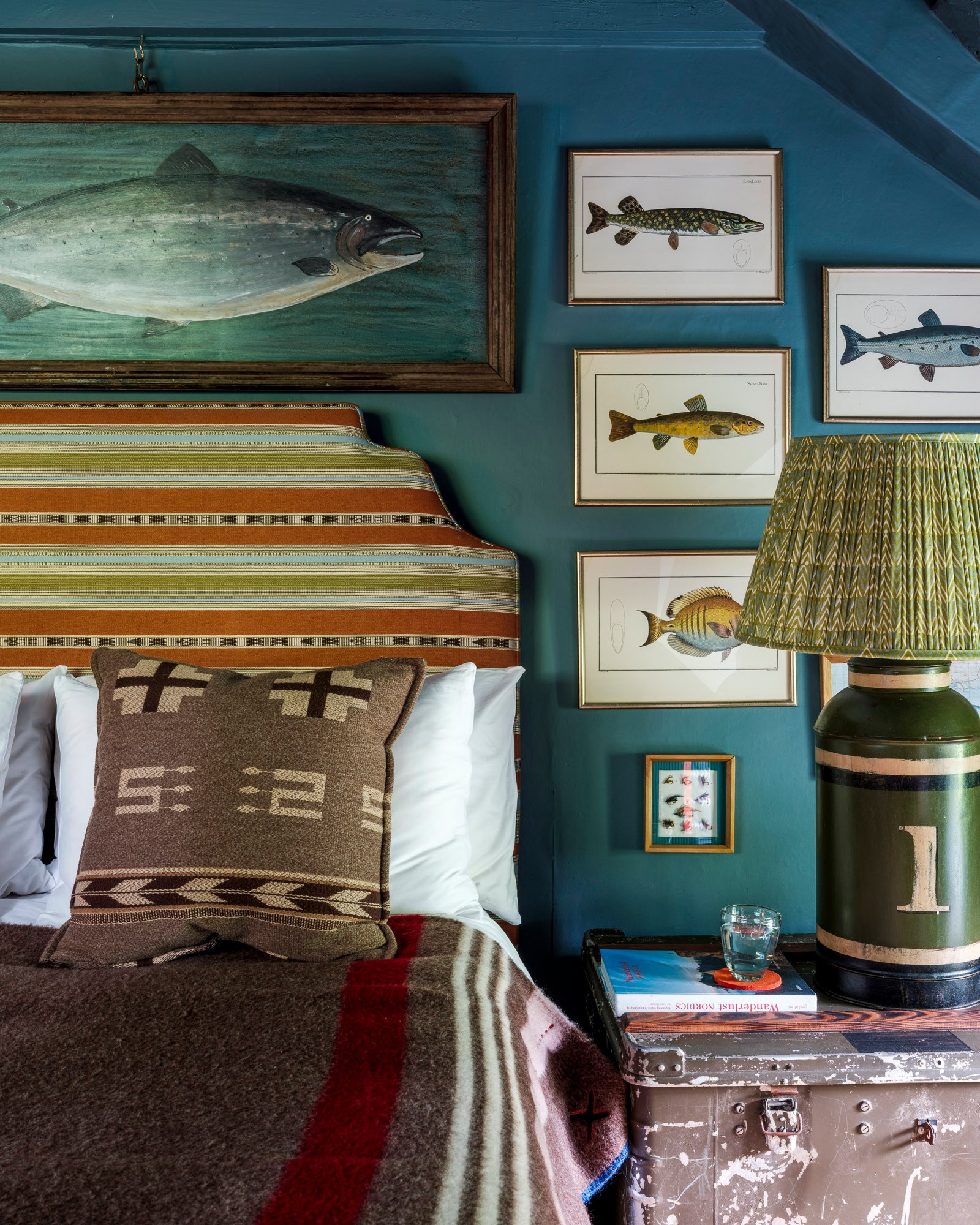 a bedroom with teal blbue wall, a collection of fish artwork on the wall, a bed with an orange and green stripe headboard and a large green pleated table lamp next to the bed