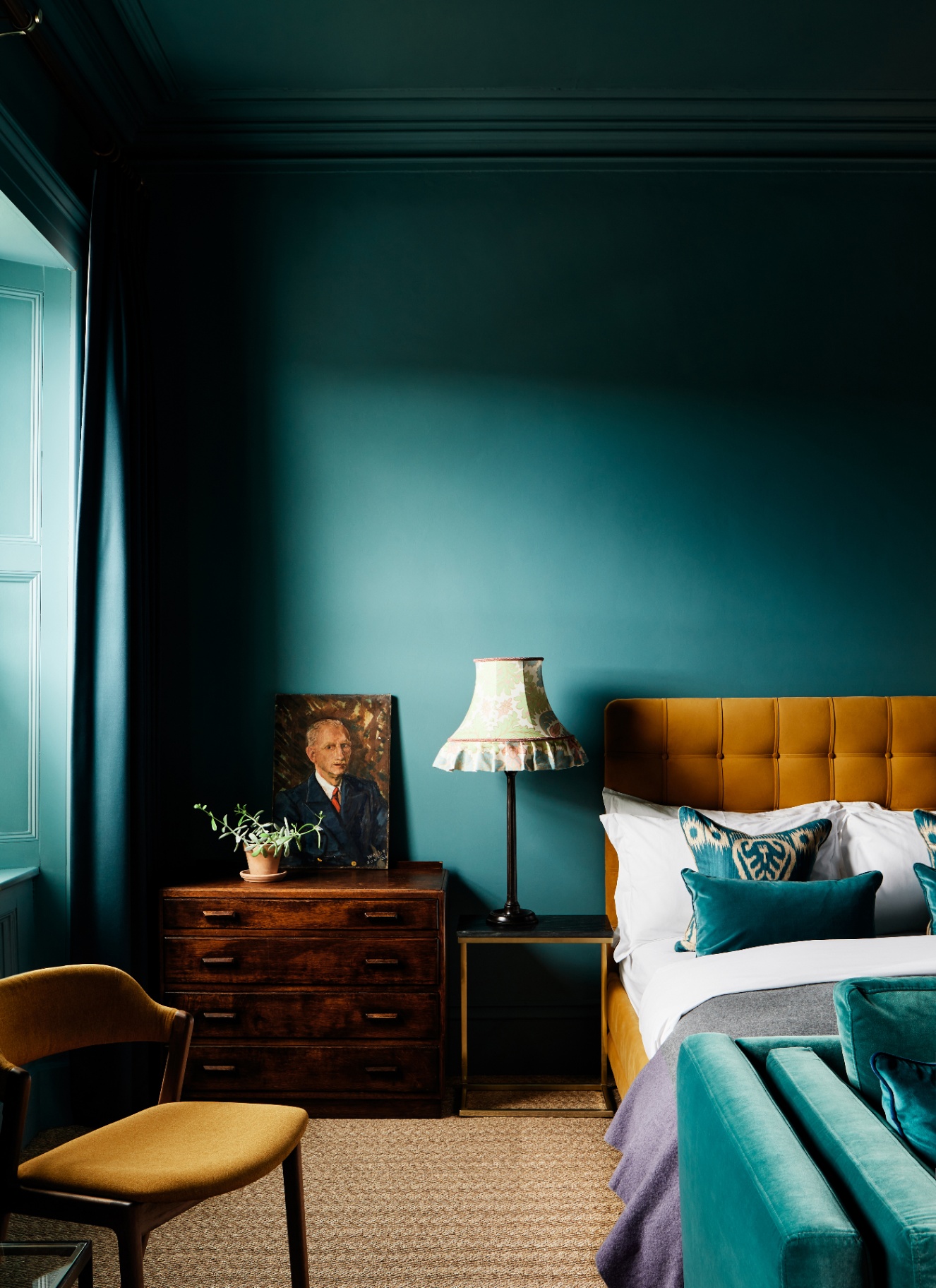 a bedroom with teal green walls and ceiling, a bed with a mustard velvet headboard and a wooden antique chest of drawers with a portrait painting