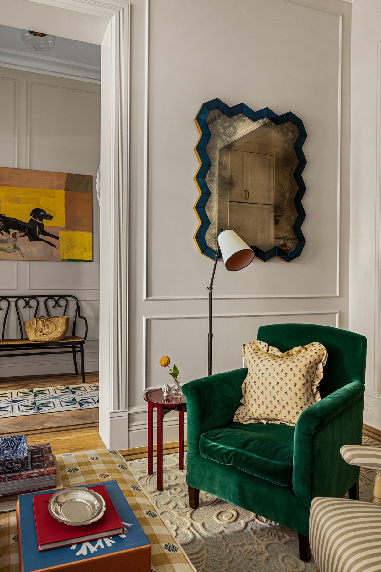 A living room with a green velvet armchair with a yellow cushion, a small circular side table and floor lamp. On the panelled wall hangs a zigzag mirror with foxed glass. Through an open doorway a hallway can be seen with a bench and large artwork of a dog