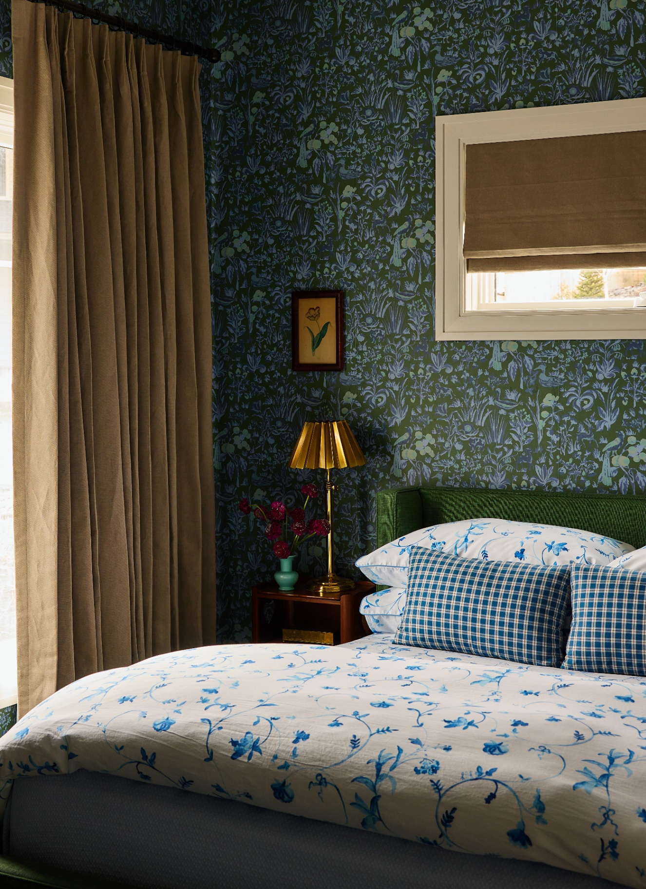 Bedroom featuring dark green botanical wallpaper, a green upholstered headboard, blue floral and gingham bed linens, warm beige drapes, and a brass table lamp