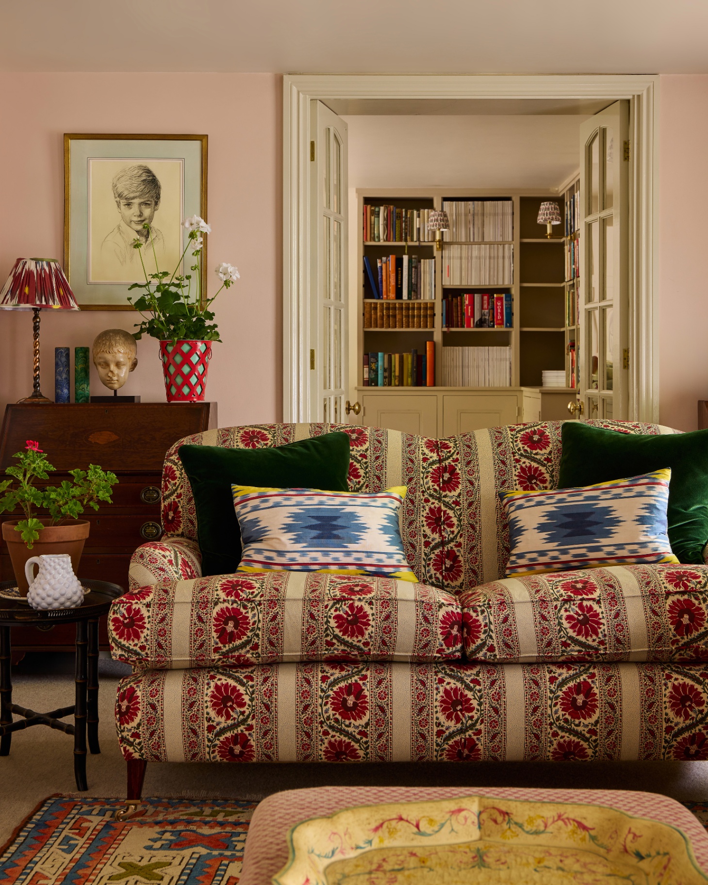a living room with a red and beige floral sofa with two green velvet cushions and two ikat cushions, with a plaster pink wall and an open door looking through to a library