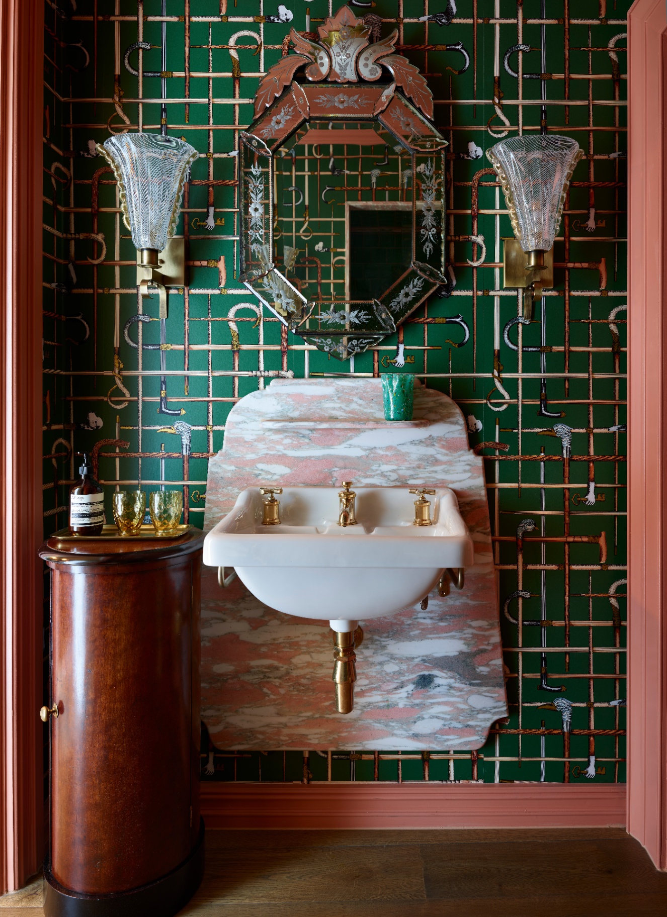 close up of a bathroom with green patterned wallpaper, a sink with apink marble splashback, a silver ornate mirror and two gold and glass sconces either side