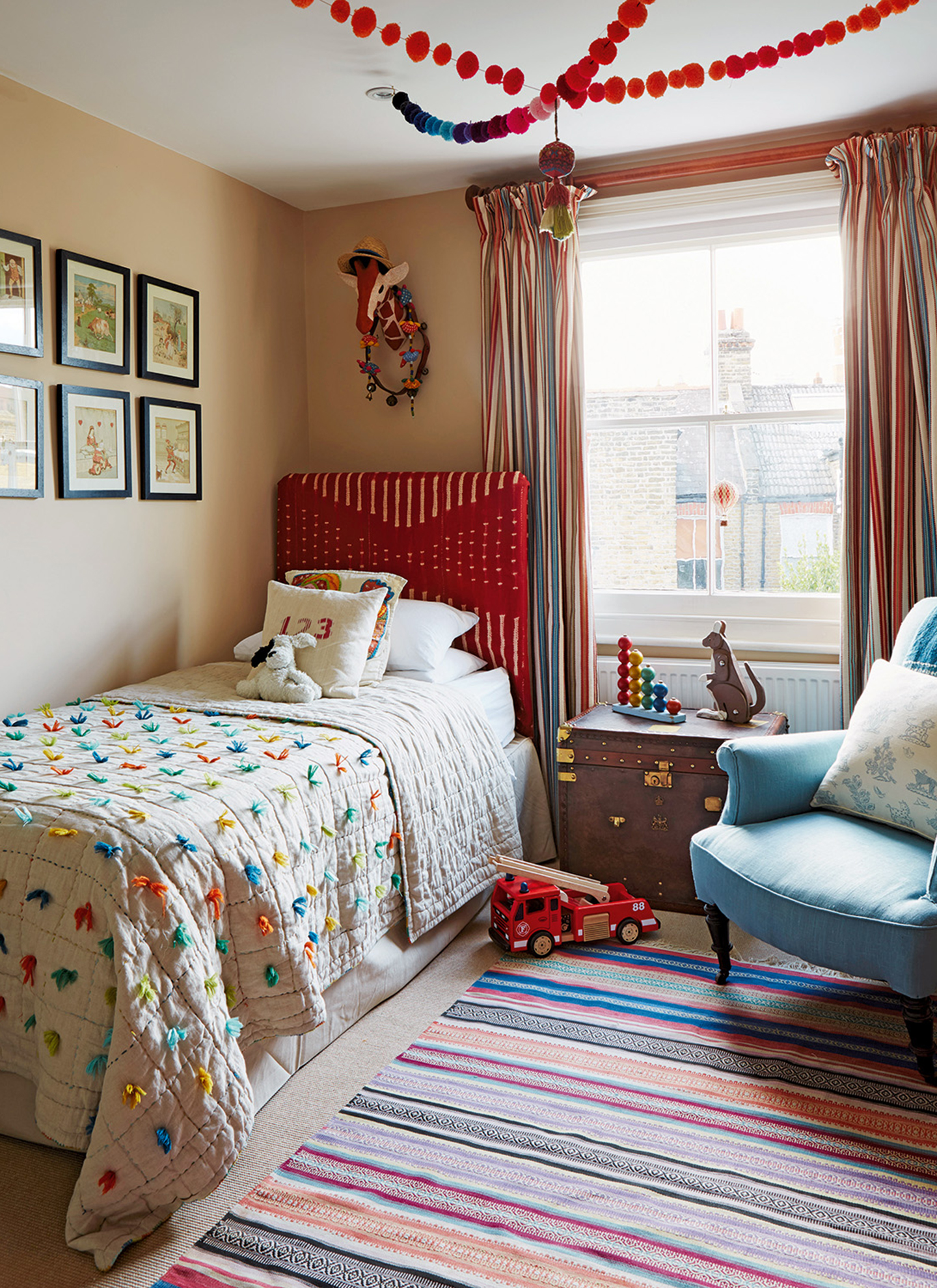 a childrens bedroom with pom pom bunting on the ceiling, a single bed with a red headboard and a vintage trunk as the bedside table