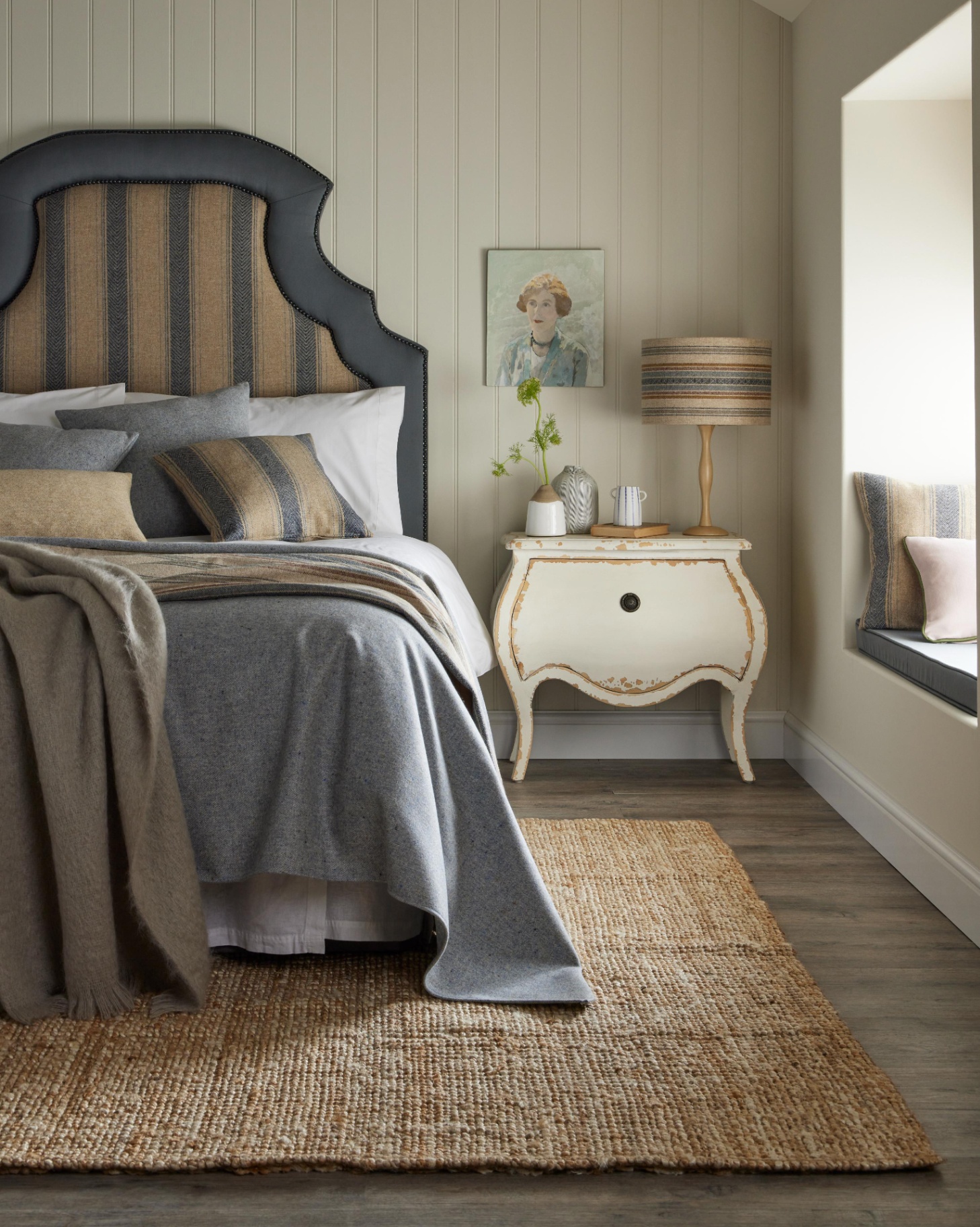 a bedroom with a jute rug, a bed with a large beige and blue striped headboard and blue and white bedding, a shappy chic white bedside table with a striped lamp